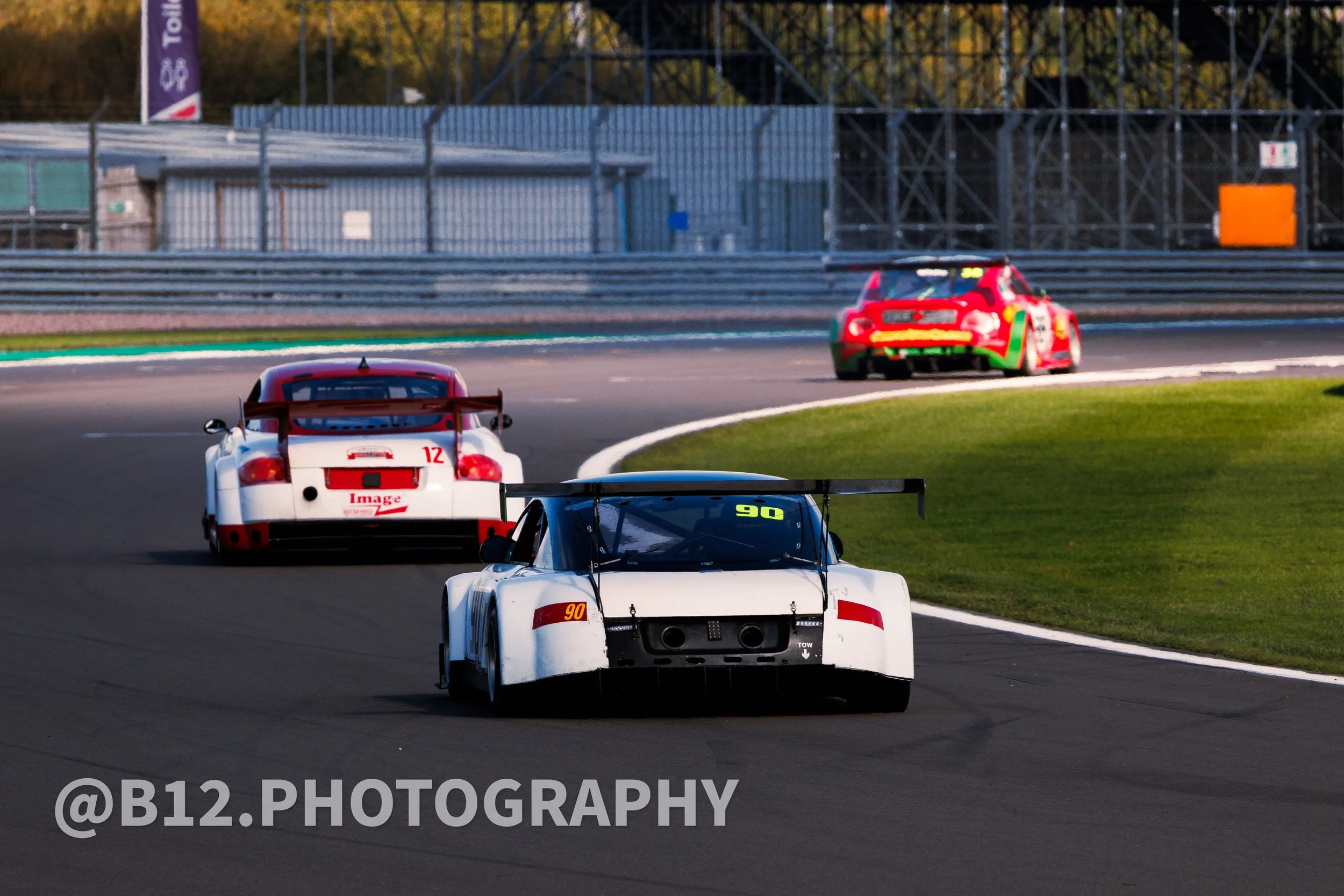 Three racing cars on a race track during a race, with a barrier and grandstands in the background.