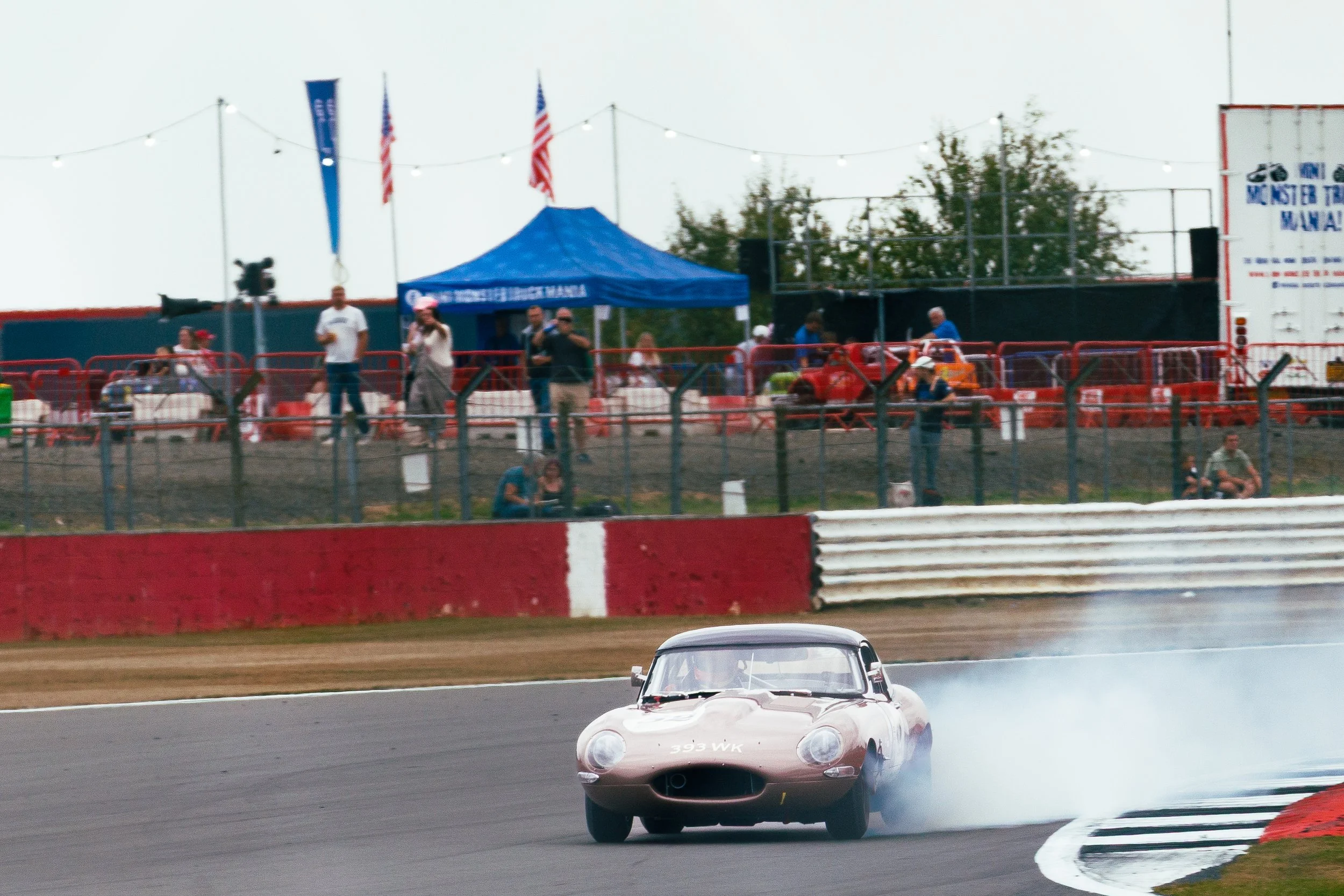 A vintage race car on a racetrack producing tire smoke during a race, with spectators and officials watching from the stands and a blue tent in the background.