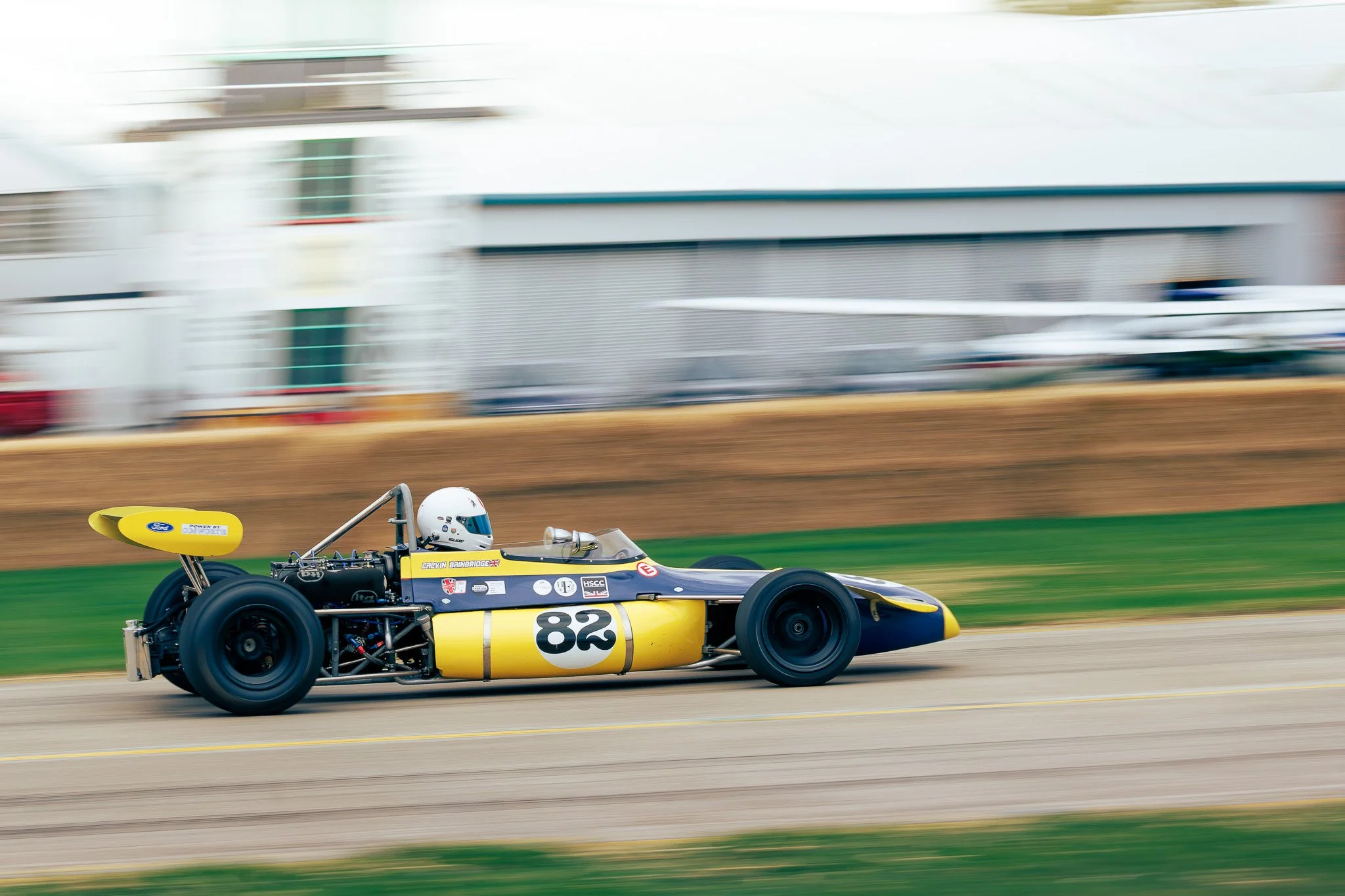 A vintage yellow and blue race car with the number 82, driven by a helmeted driver, speeds along a race track with a blurred background of buildings and fences.