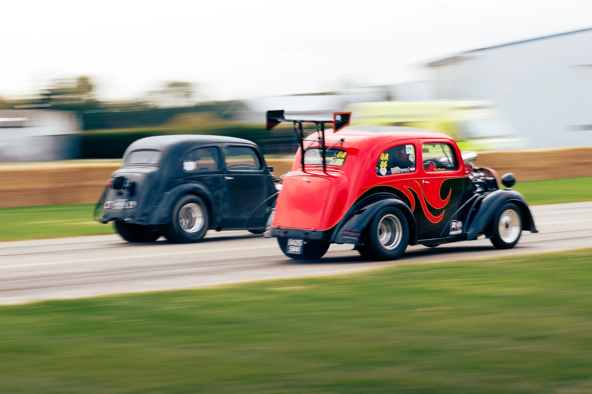 Two vintage race cars on a track, one black and one red with black and yellow flame designs, speeding past each other in a race.