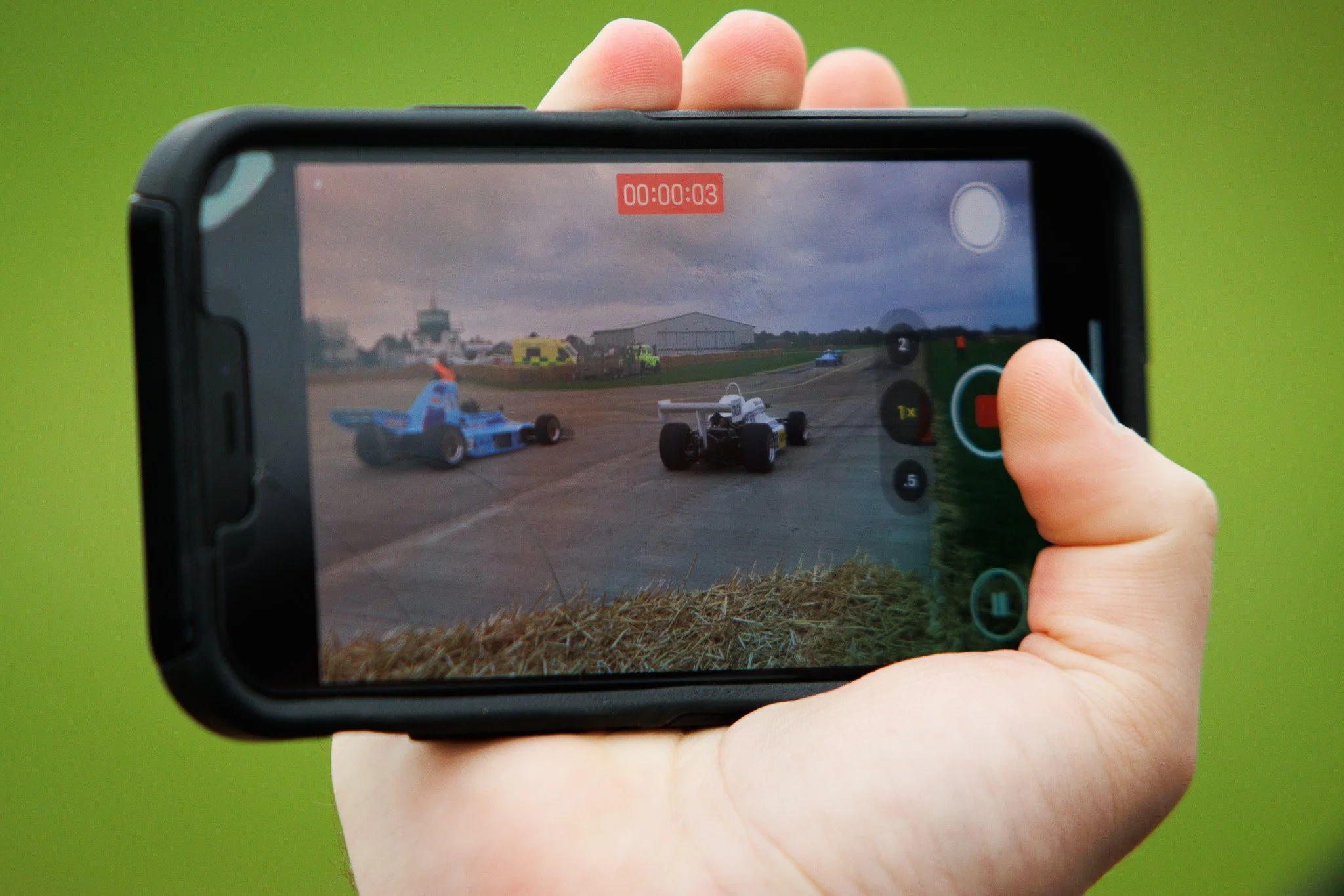 A person holding a smartphone recording a race car crash on a race track. The phone's screen shows multiple race cars, with one white car colliding with a blue car, under a cloudy sky.