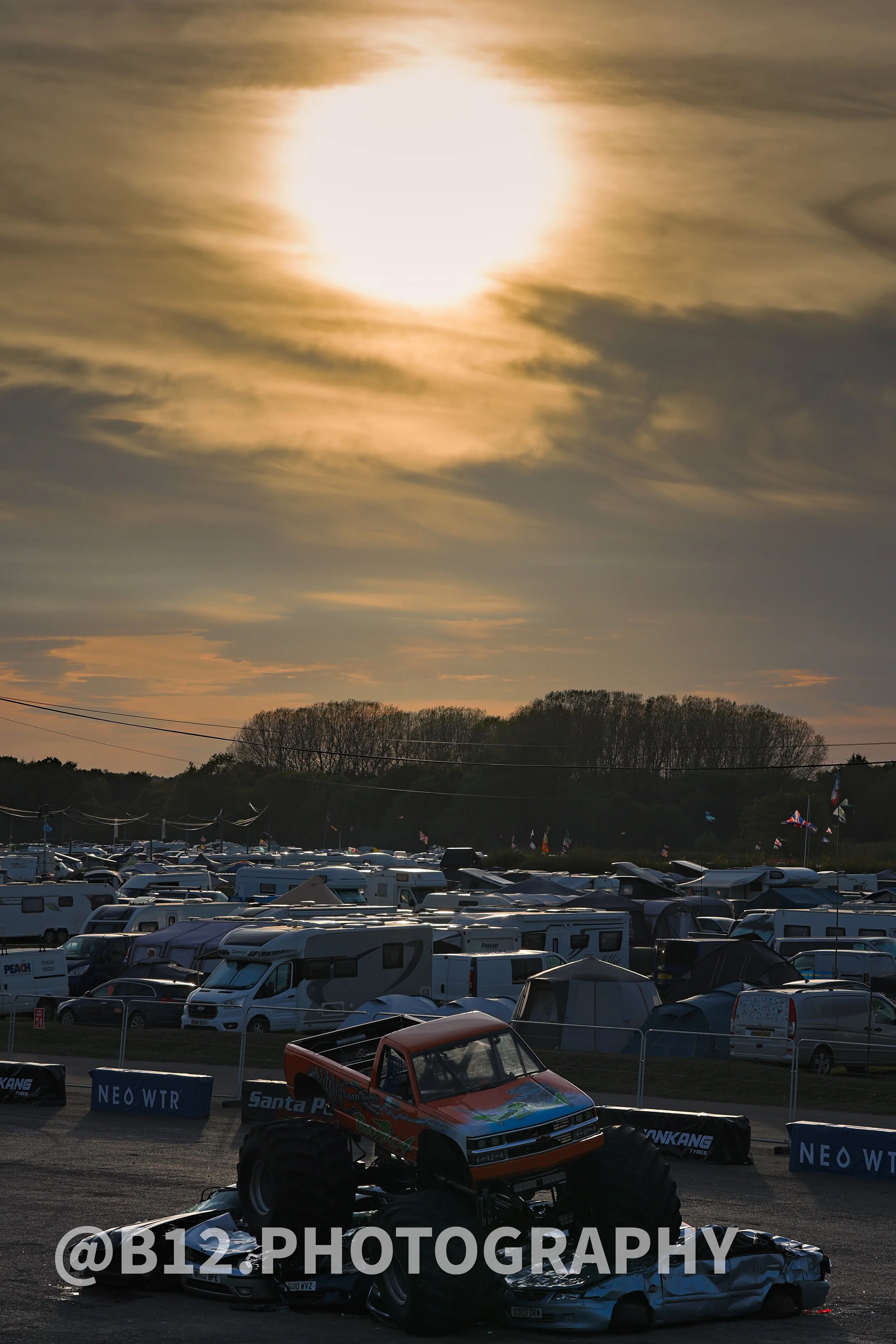 A scene at a motorsports event featuring a monster truck parked on top of a damage sports car, with a large RV parking lot in the background under a cloudy sky over the sunset.