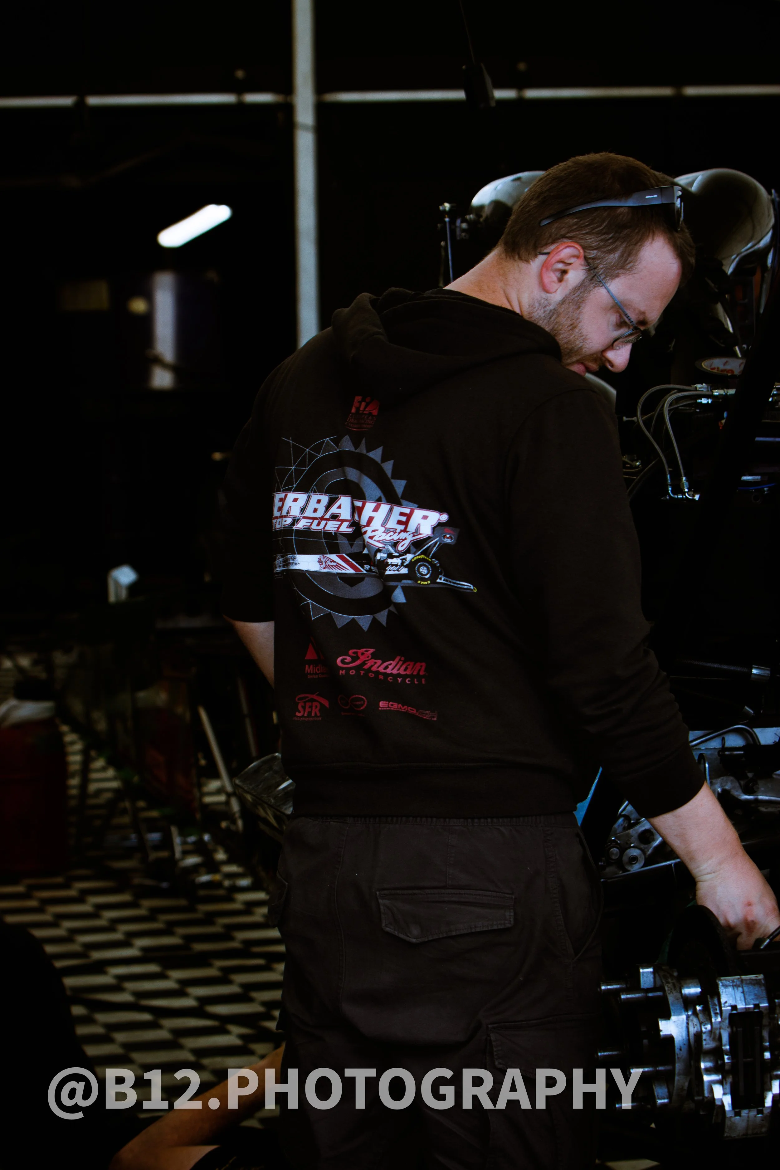 A man wearing glasses and a black hoodie with racing logos, working on a motorcycle in a garage with checkered floor.