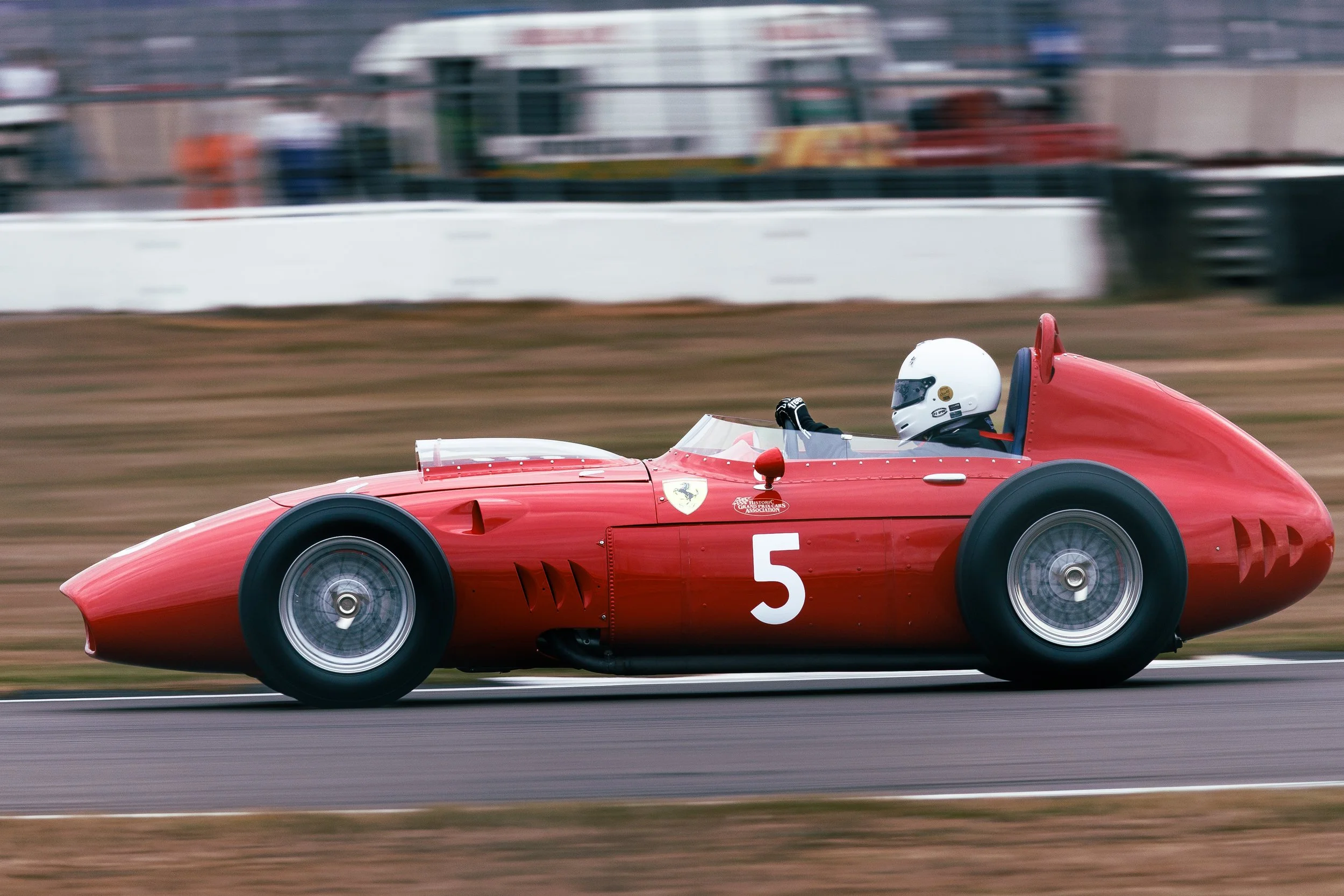 A vintage red race car with the number 5 on the side speeding on a race track, with a driver in a white helmet visible.