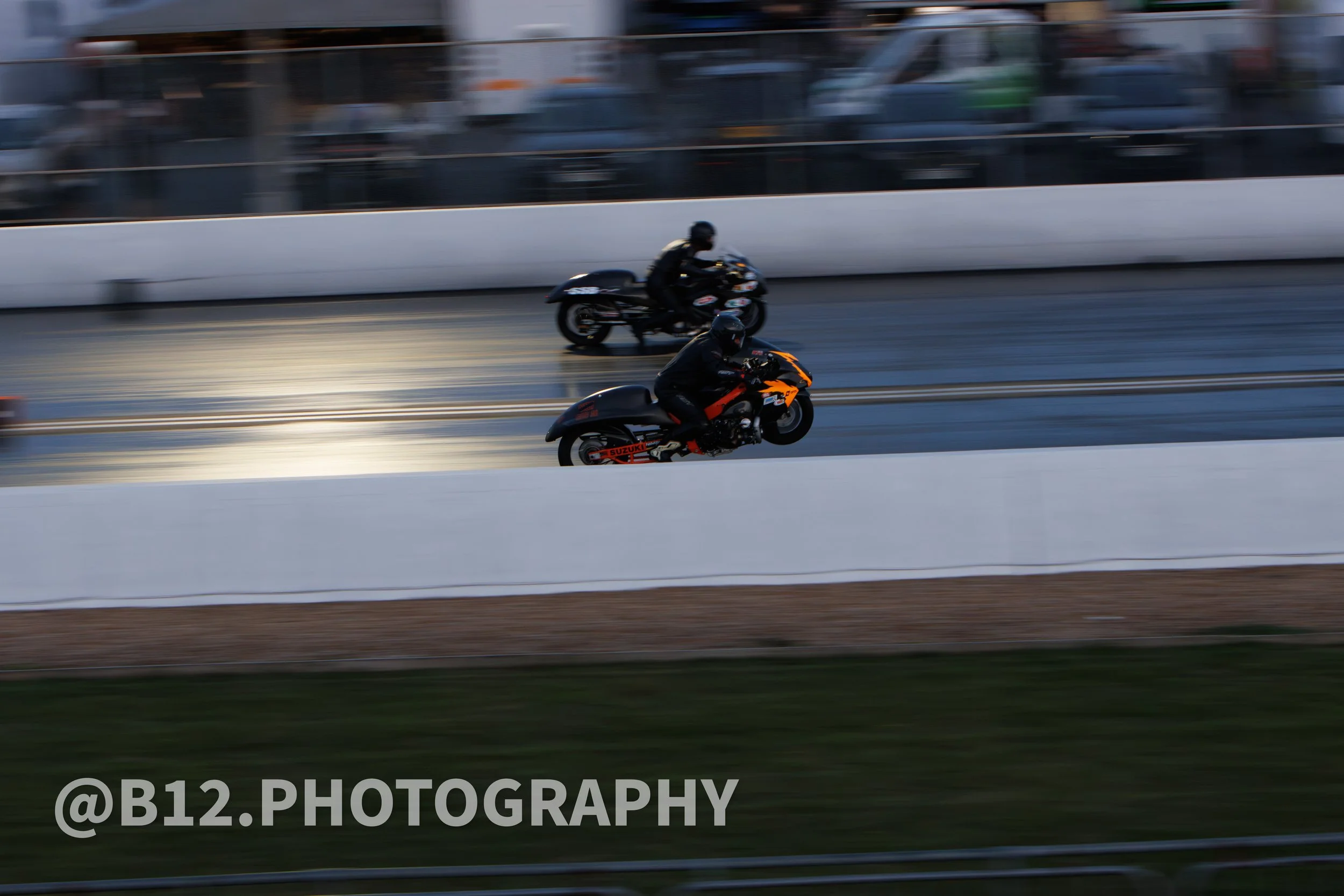 Two motorcyclists racing on a drag strip, one in orange and black gear, the other in black gear, with blurred background indicating high speed.