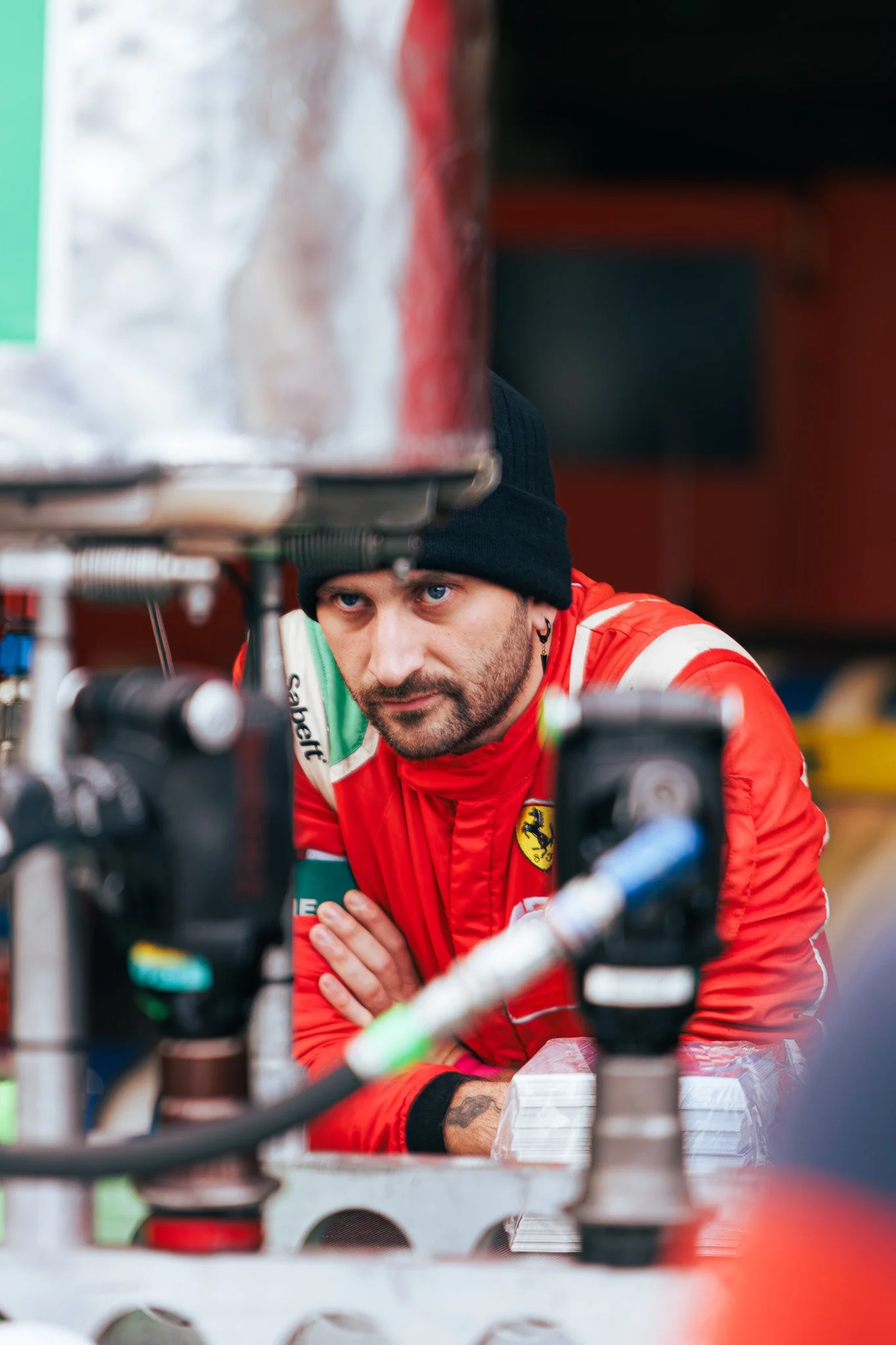 A man in a red racing suit with Ferrari branding, wearing a black beanie, looking intently through equipment in a garage or workshop.