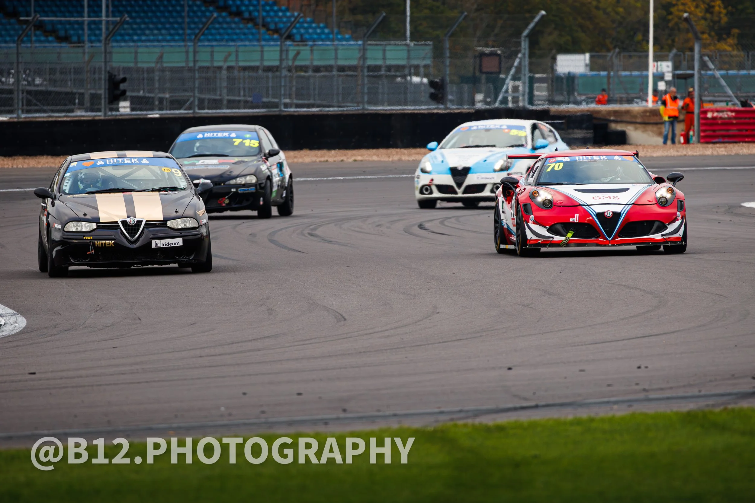 Race cars on a racetrack, with a black, gray, white, and red cars in passing, and some officials or spectators watching in the background.