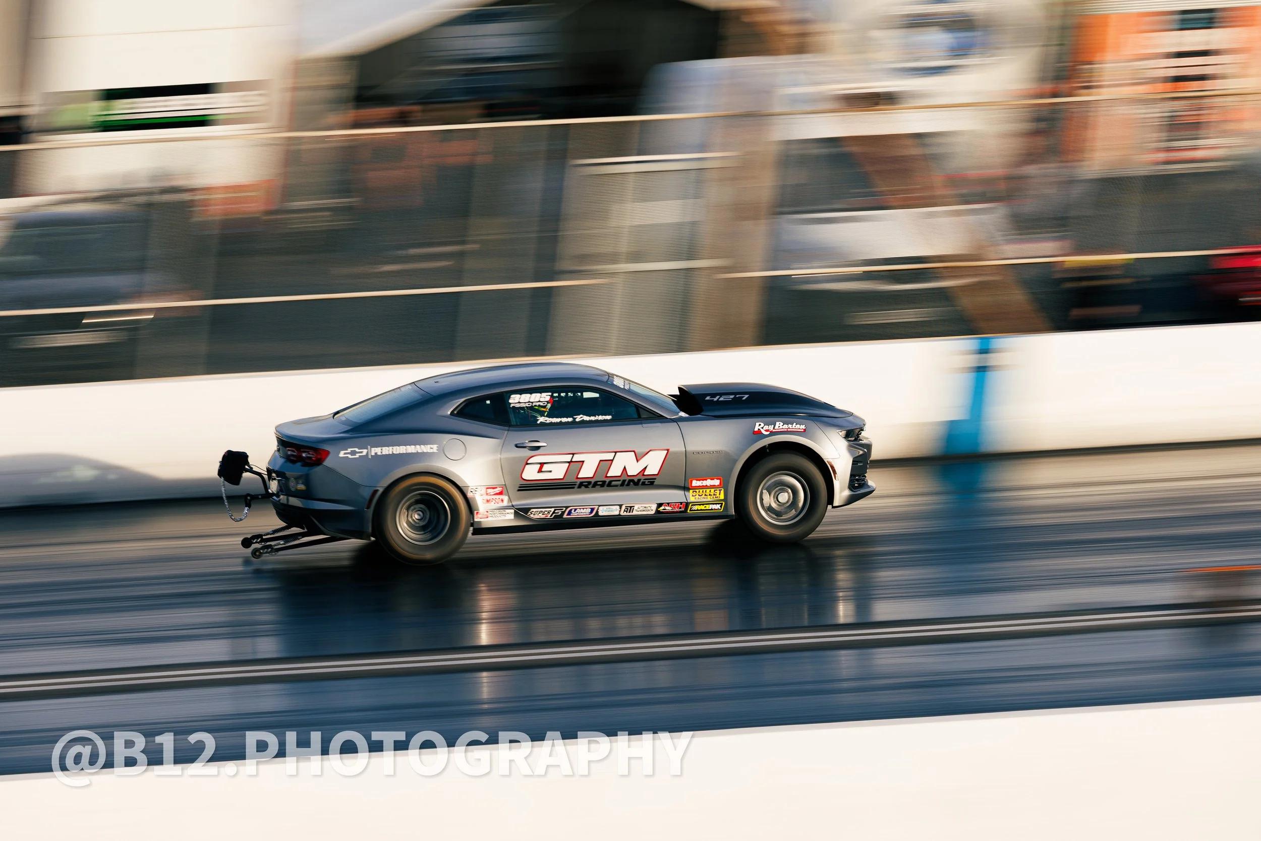 A silver race car with GTM Racing and Chevrolet Performance decals speeding down a drag strip, with slight motion blur and a blurred background.