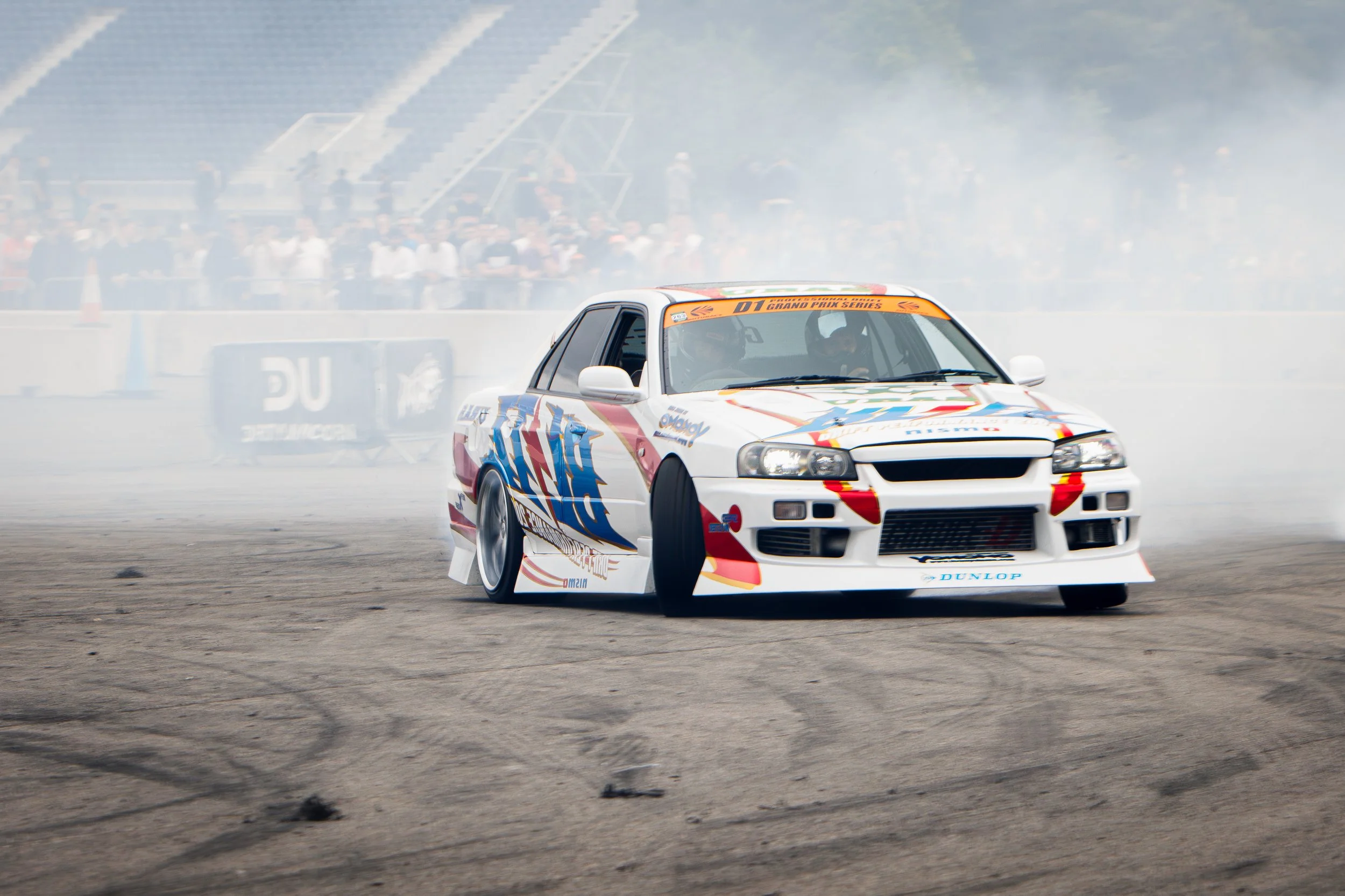A race car performing a drift maneuver on the track with smoke trailing behind, surrounded by spectators in the stands.