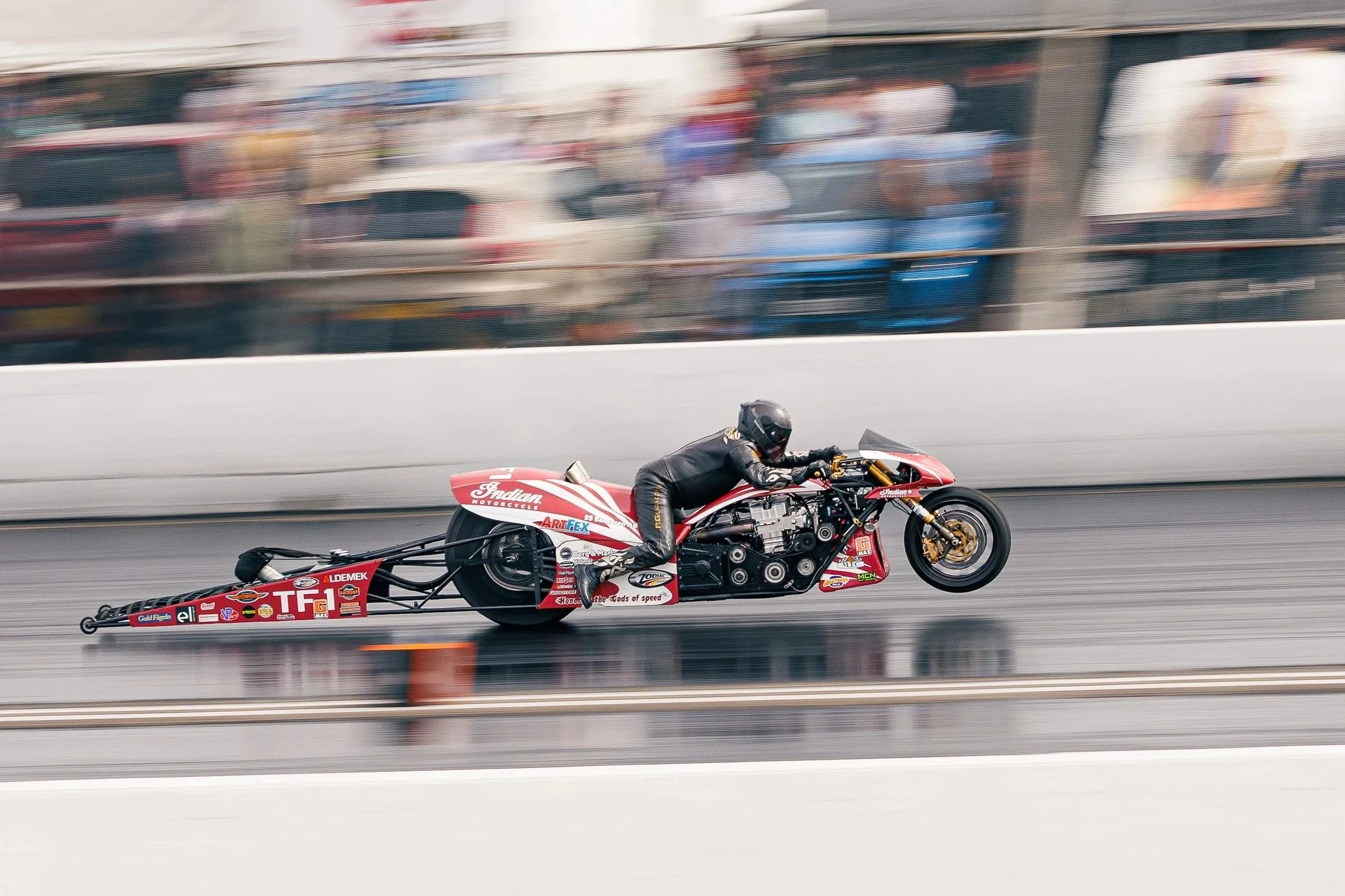 A nitro drag race motorcycle with a rider in black gear and helmet speeding on the track, with colorful blurred spectators and vehicles in the background.