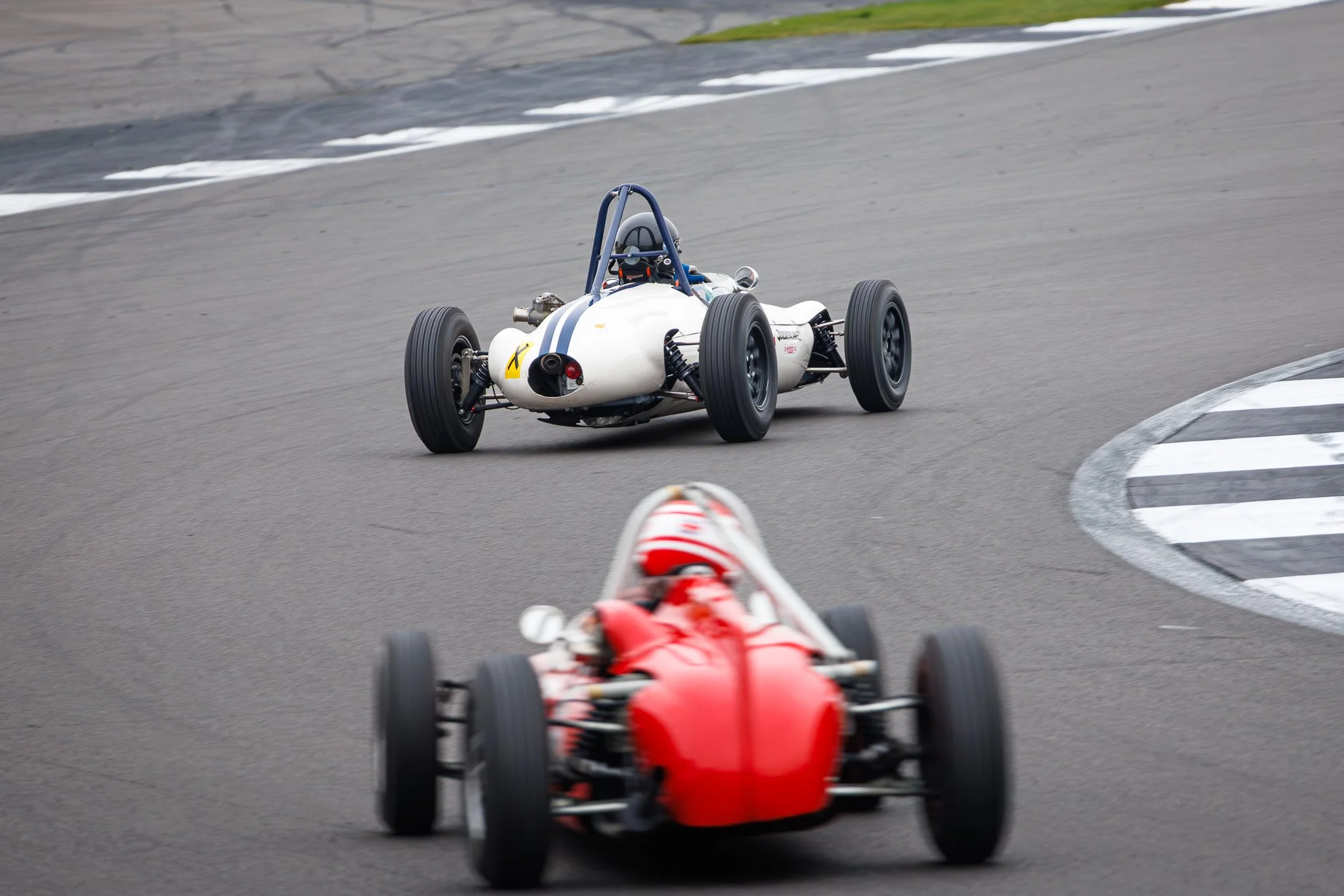 Two vintage race cars on a race track, with one red car in the foreground and a white car with blue stripes in the background, navigating a turn.