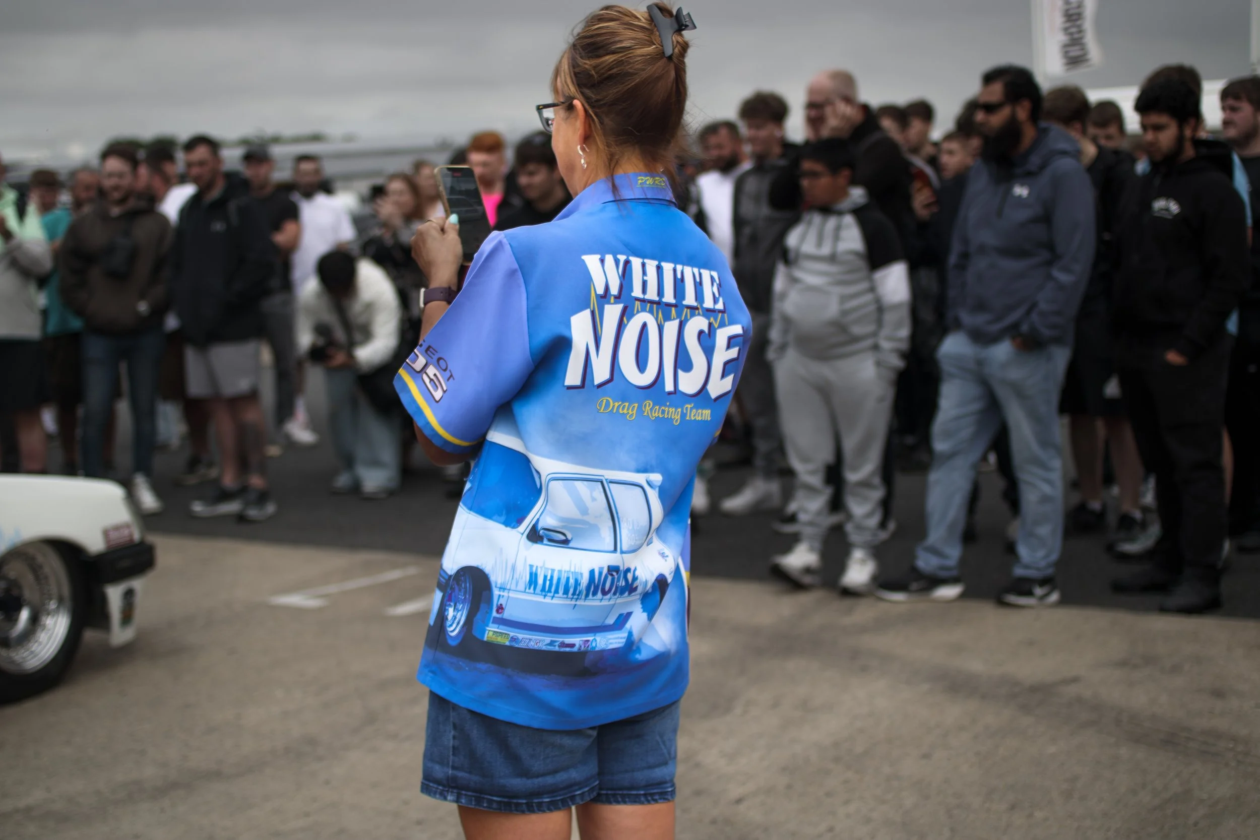 A woman with glasses and a blue shirt, with 'WHITE NOISE Drag Racing Team' written on it, is taking a photo or video with her phone in front of a group of people at a race event.