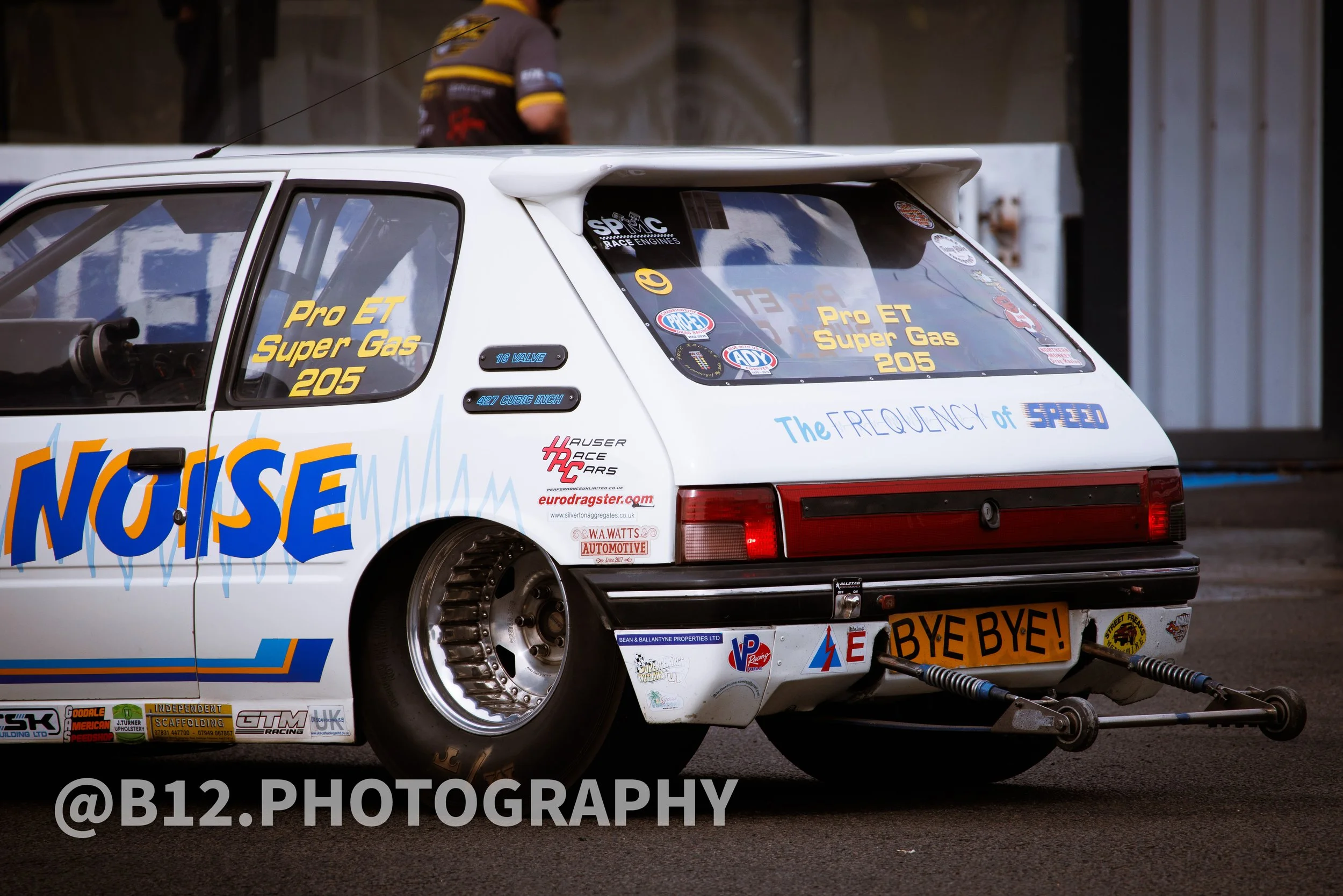 A modified racing hatchback car with large rear tires, graphics, stickers, and the phrase 'The FREQUENCY of SPEED' on the rear window, parked at a racing event.