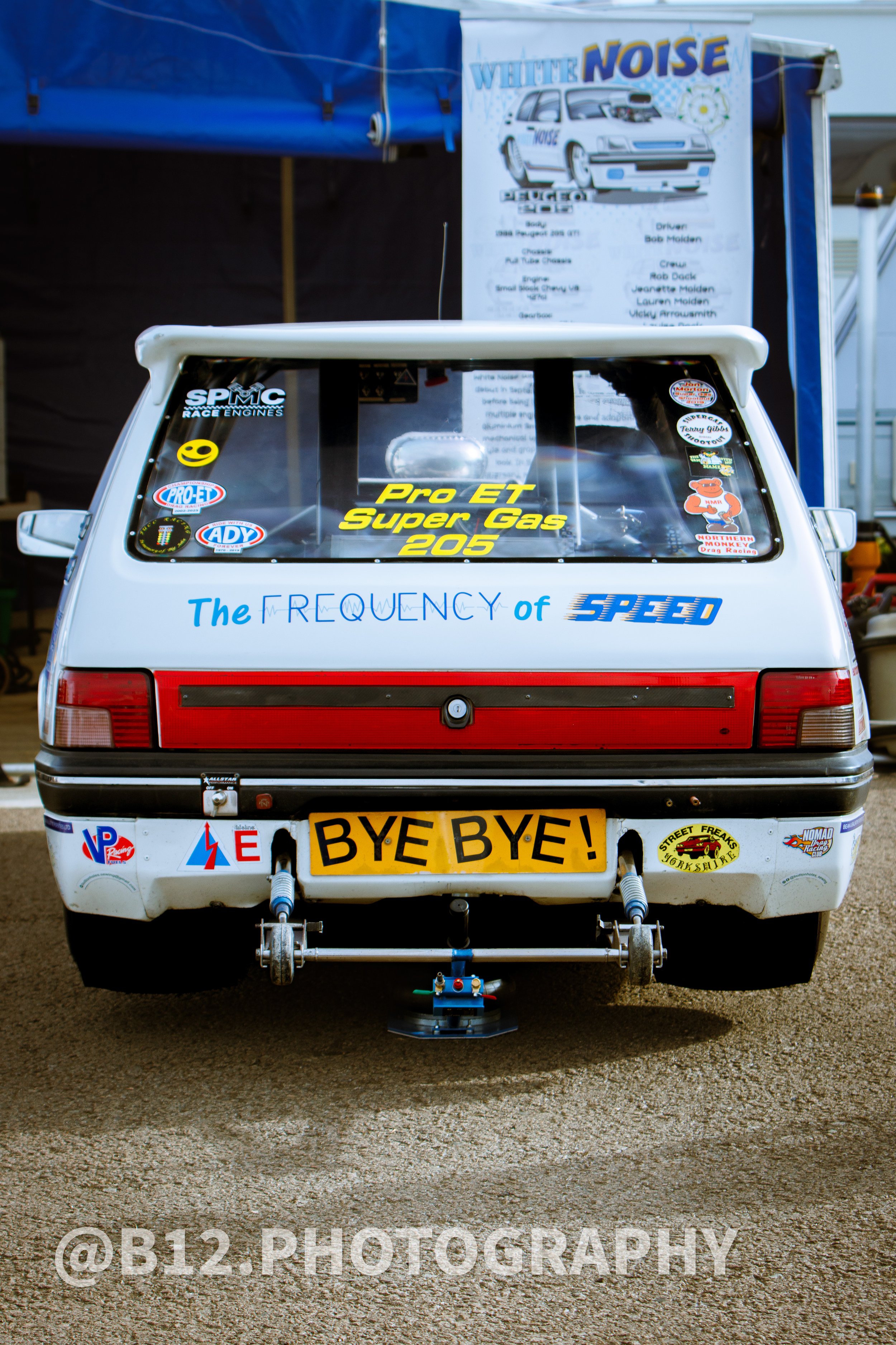 Rear view of a white race car with various stickers and a license plate reading 'BYE BYE!' in yellow and black. The car has a large rear wing and a rear-wheel drive setup. The car is parked on a dirt surface with a blue tent and a poster about a raci