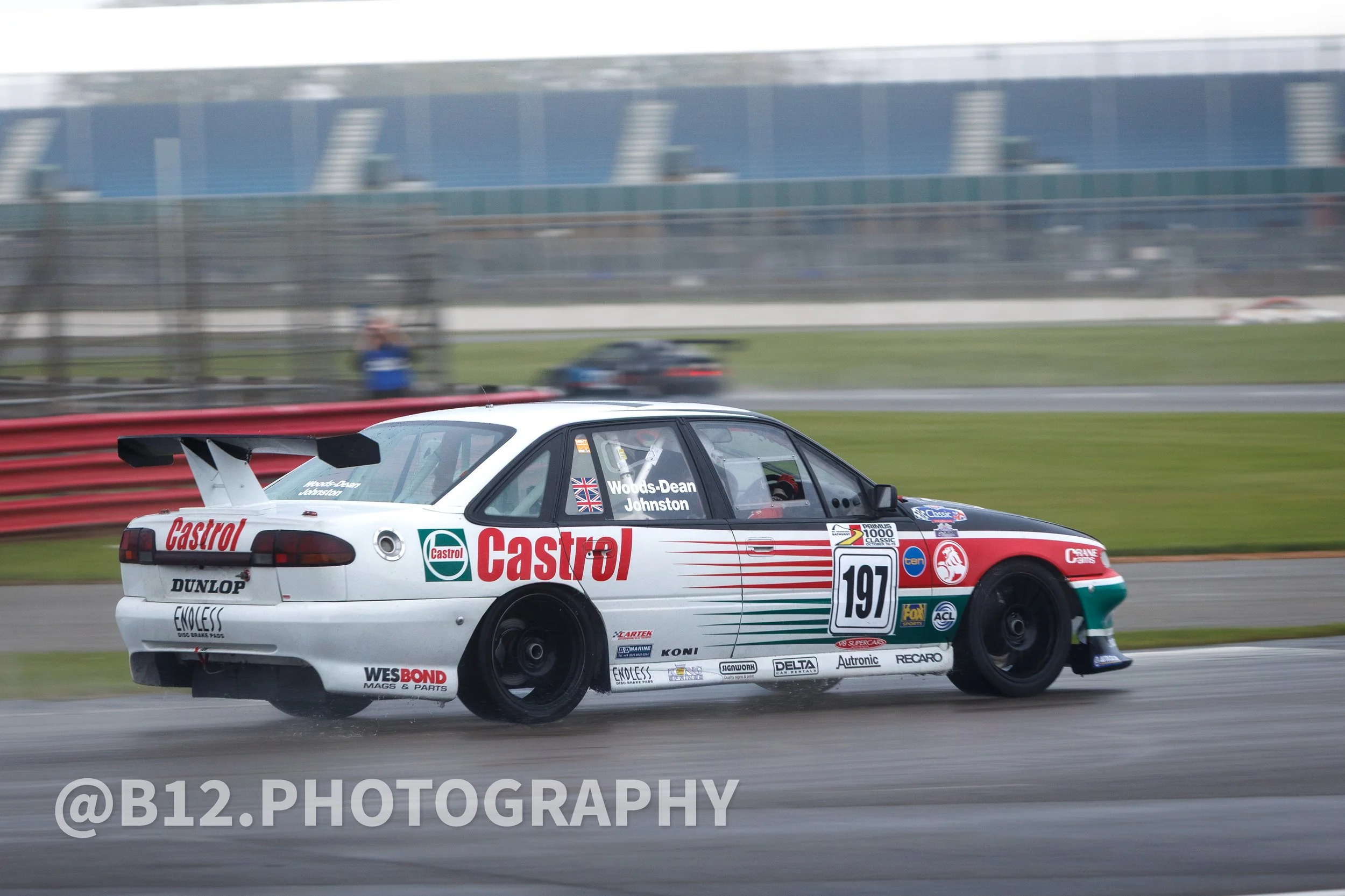 A race car with a white, red, and green livery driving on a wet race track during a storm, with a blurred background of grandstands and other vehicles.