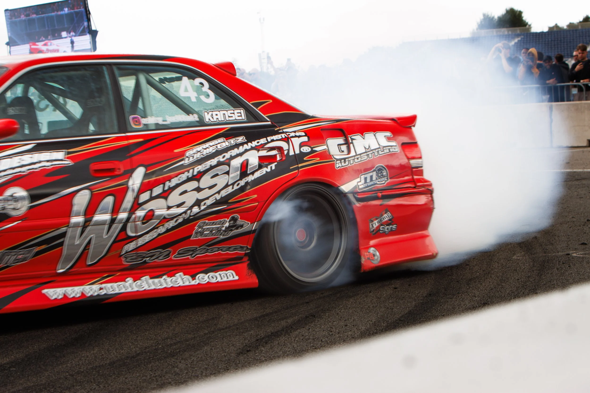 A red race car performing a burnout on a racetrack, with smoke billowing from the rear tires and a crowd watching in the background.
