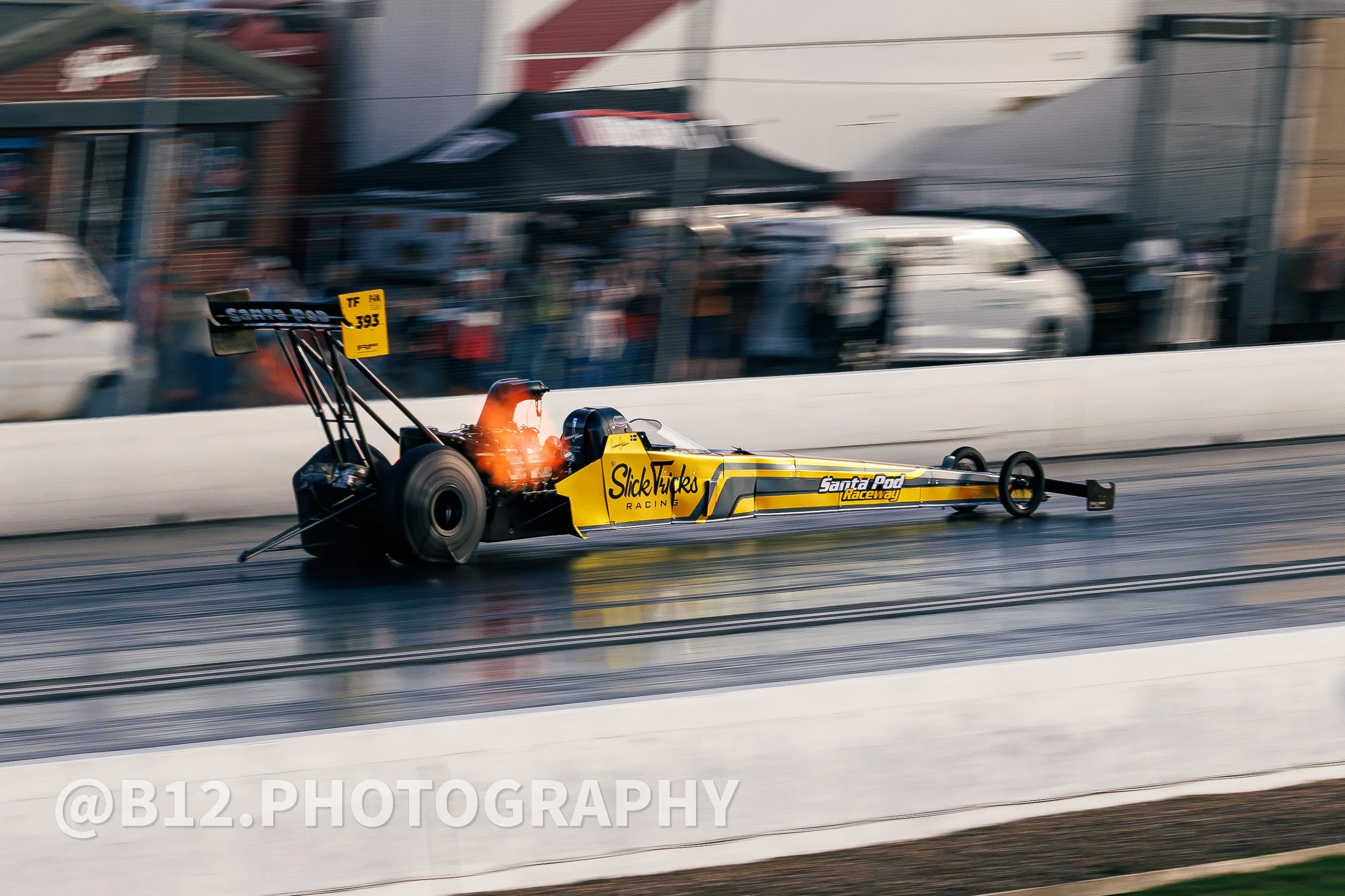 A dragster race car speeds down a track at Santa Pod Raceway, with blurred background and flames coming from the exhaust. The car is yellow with black and gray accents.