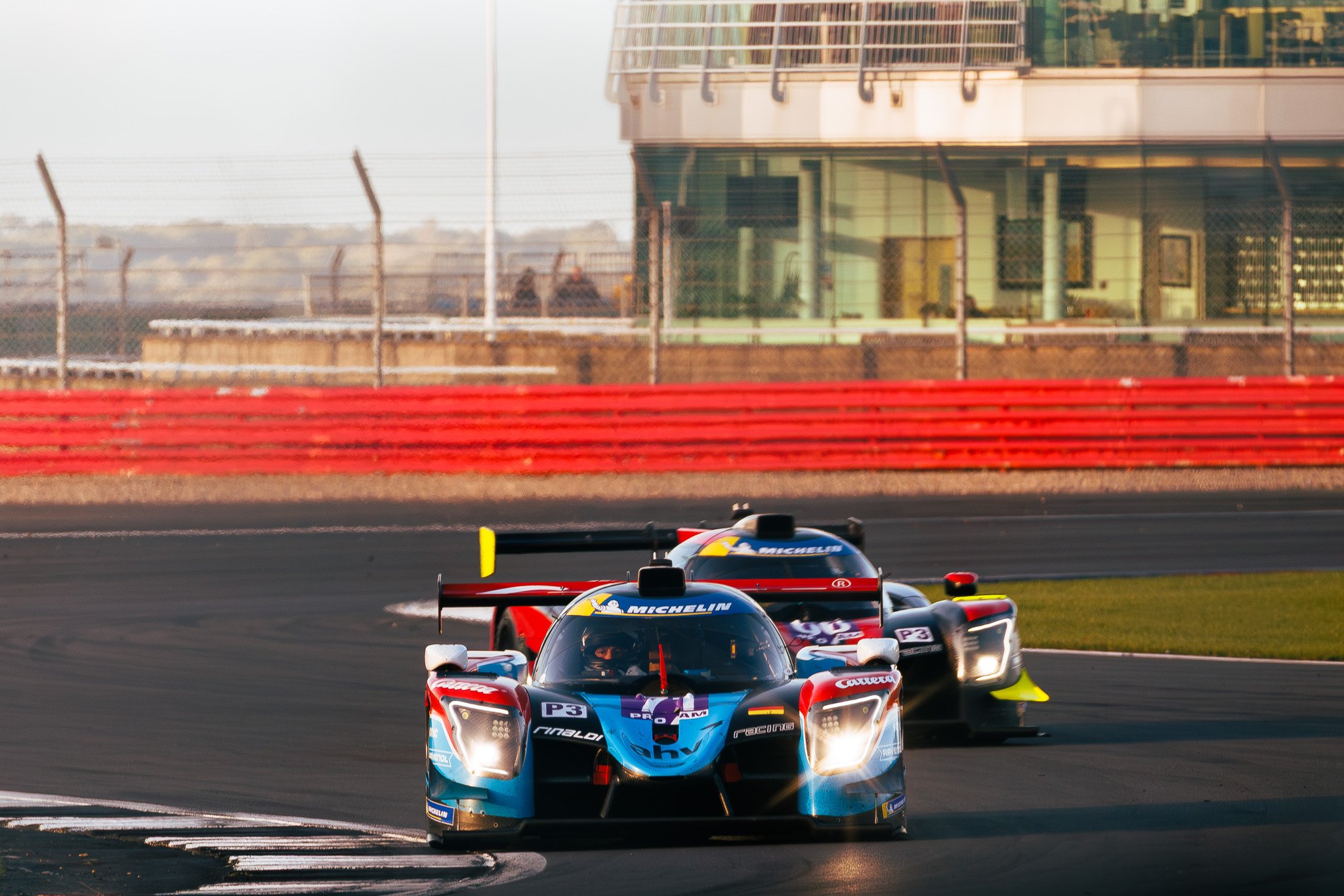 Race cars on a track during an early morning or late afternoon race, with a red barrier and building in the background.