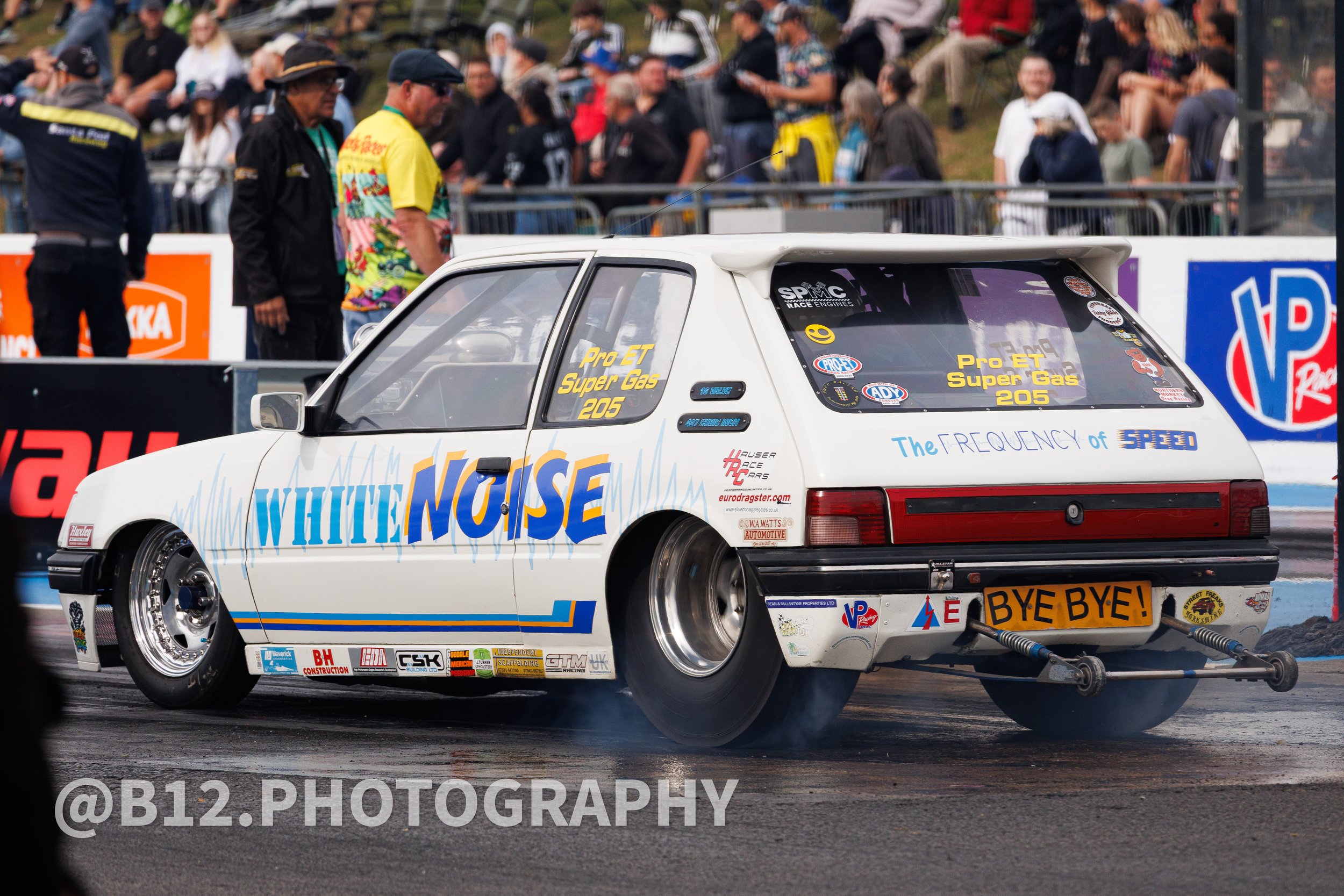A white race car with colorful decals and sponsor logos, performing a burnout at a drag strip during a racing event, with people watching in the background.