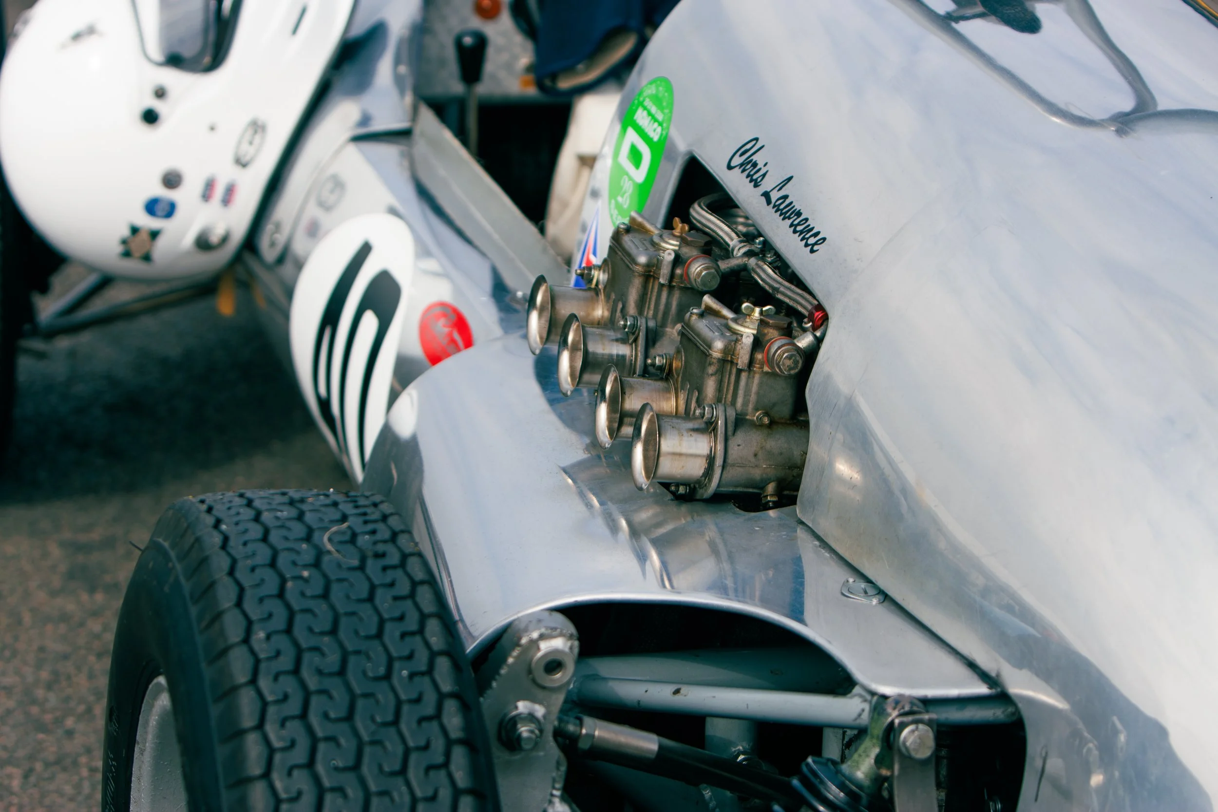 Close-up of a vintage race car's engine and front wheel, with the driver helmet visible in the background.