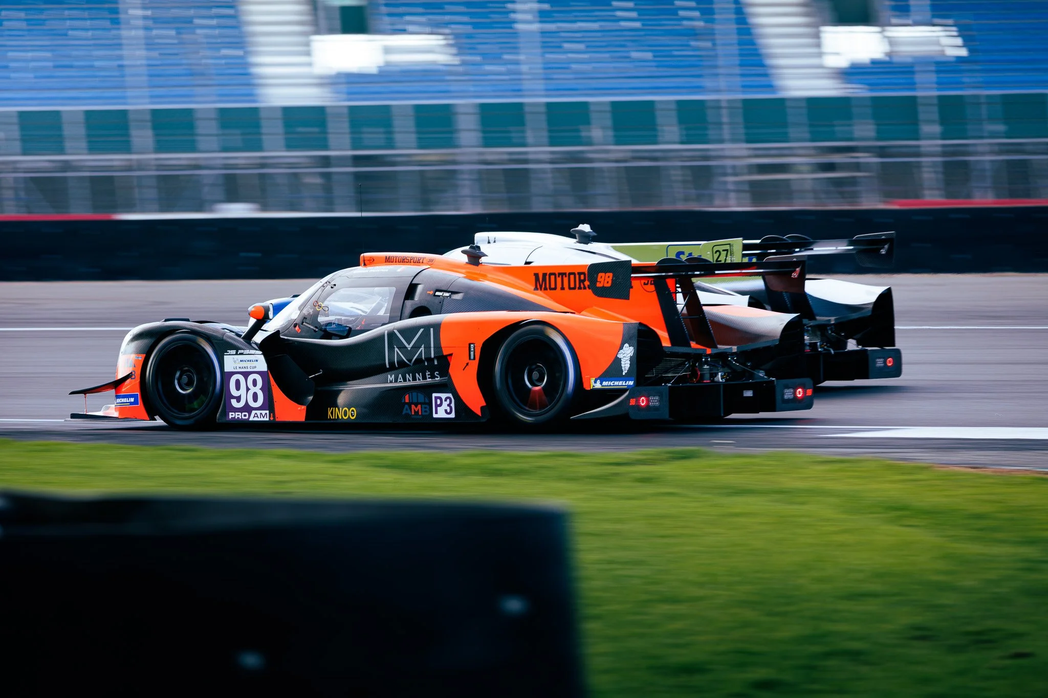A sleek race car speeding on a racetrack with blurred background, featuring black and orange colors and various sponsor logos including Michelin and Michelino.