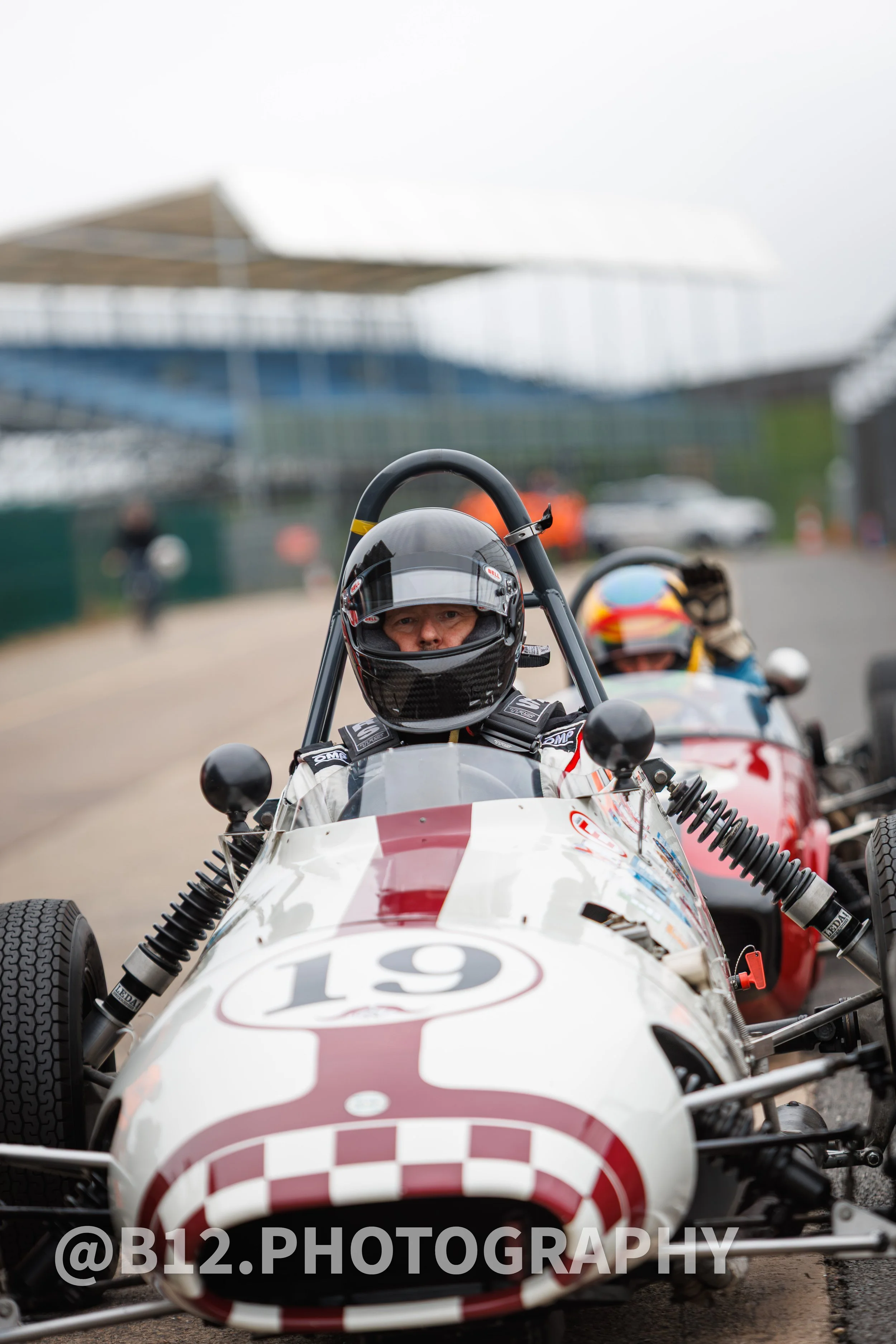 A race car driver in a helmet sitting in a vintage open-wheel race car with the number 19, at a racetrack with a blurred background of structures and other race cars.