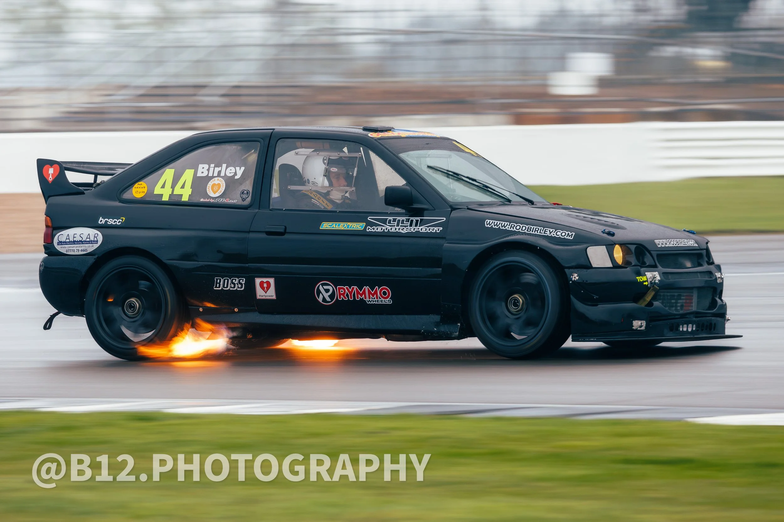 A black race car on a track with flames coming out from the rear tire area during a race.