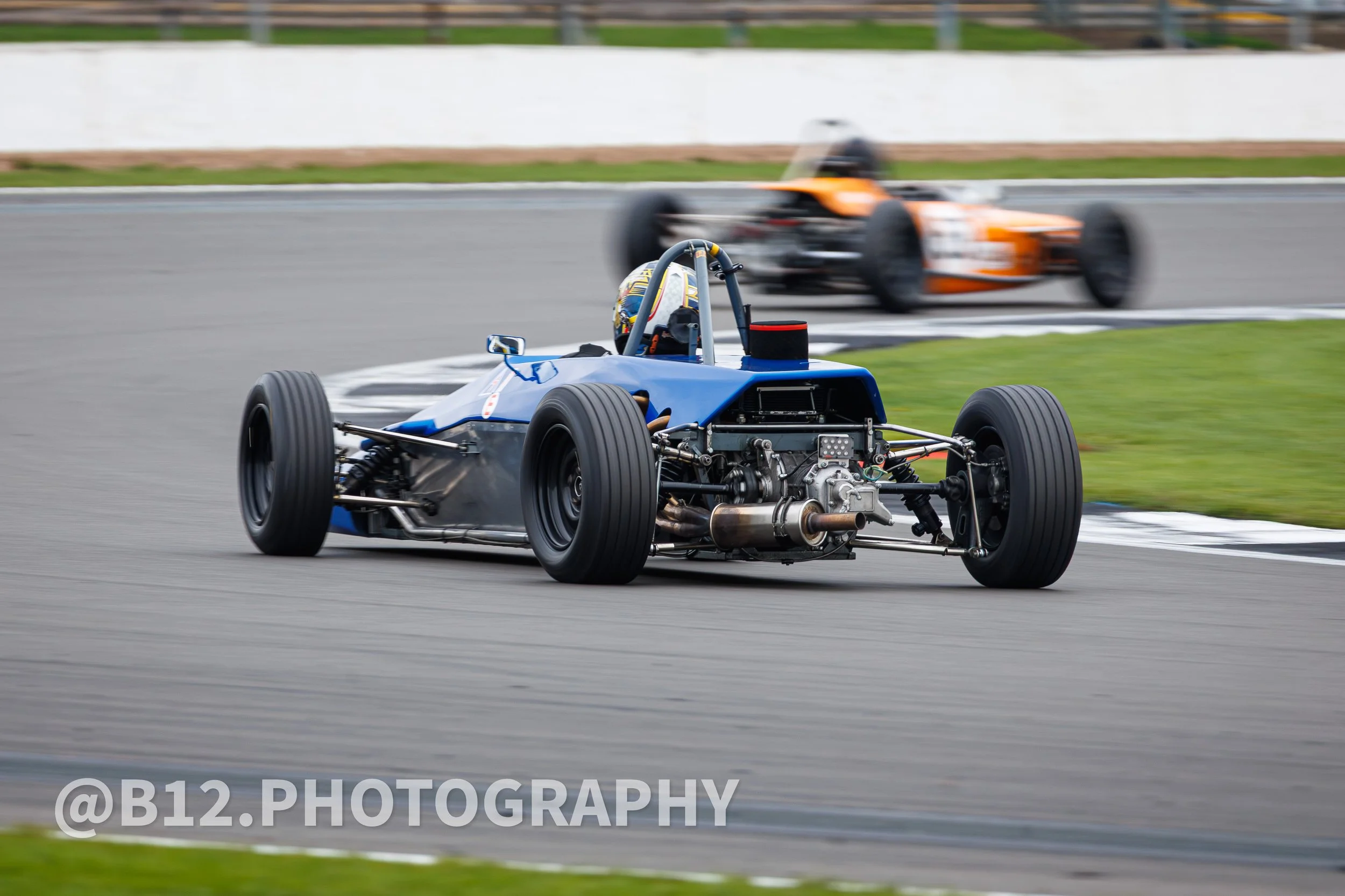 Open-wheel race car on a track with a blurred orange car in the background during a race.