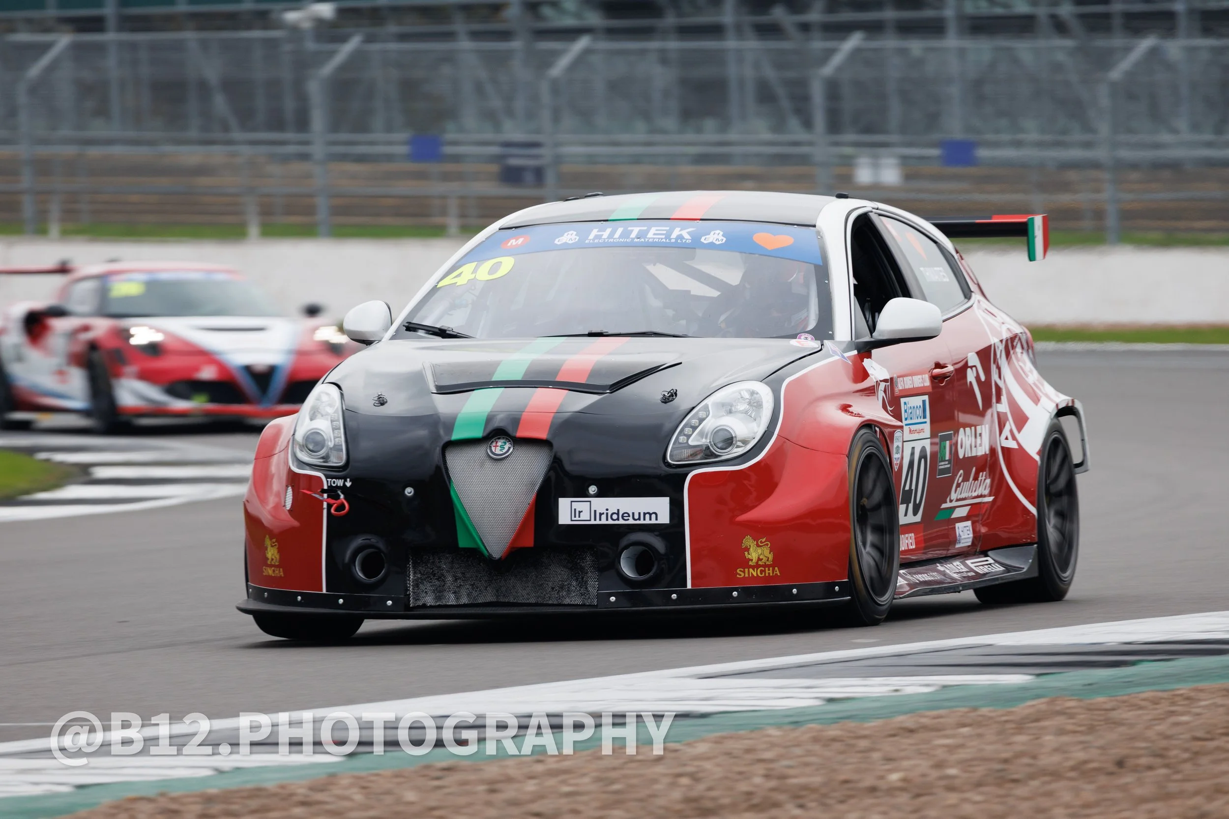 Race car on track with red, black, and green livery, leading a second red race car in the background.
