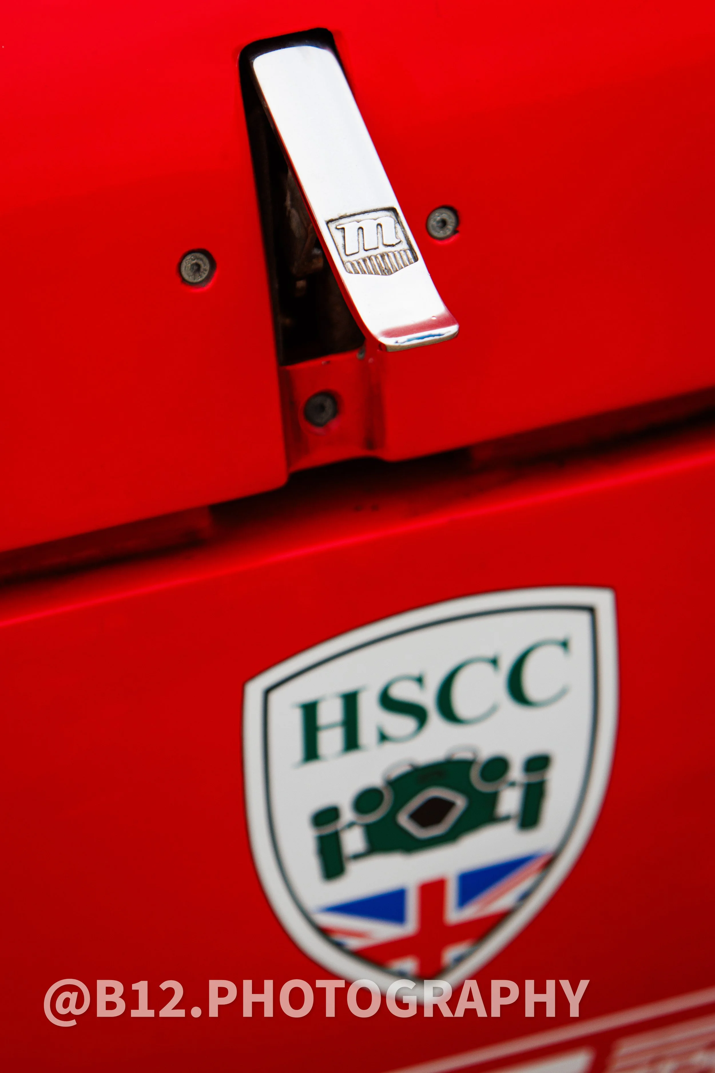 Close-up of a red metal surface with a chrome latch bearing an embossed logo, and a badge with the HSCC logo and UK flag.