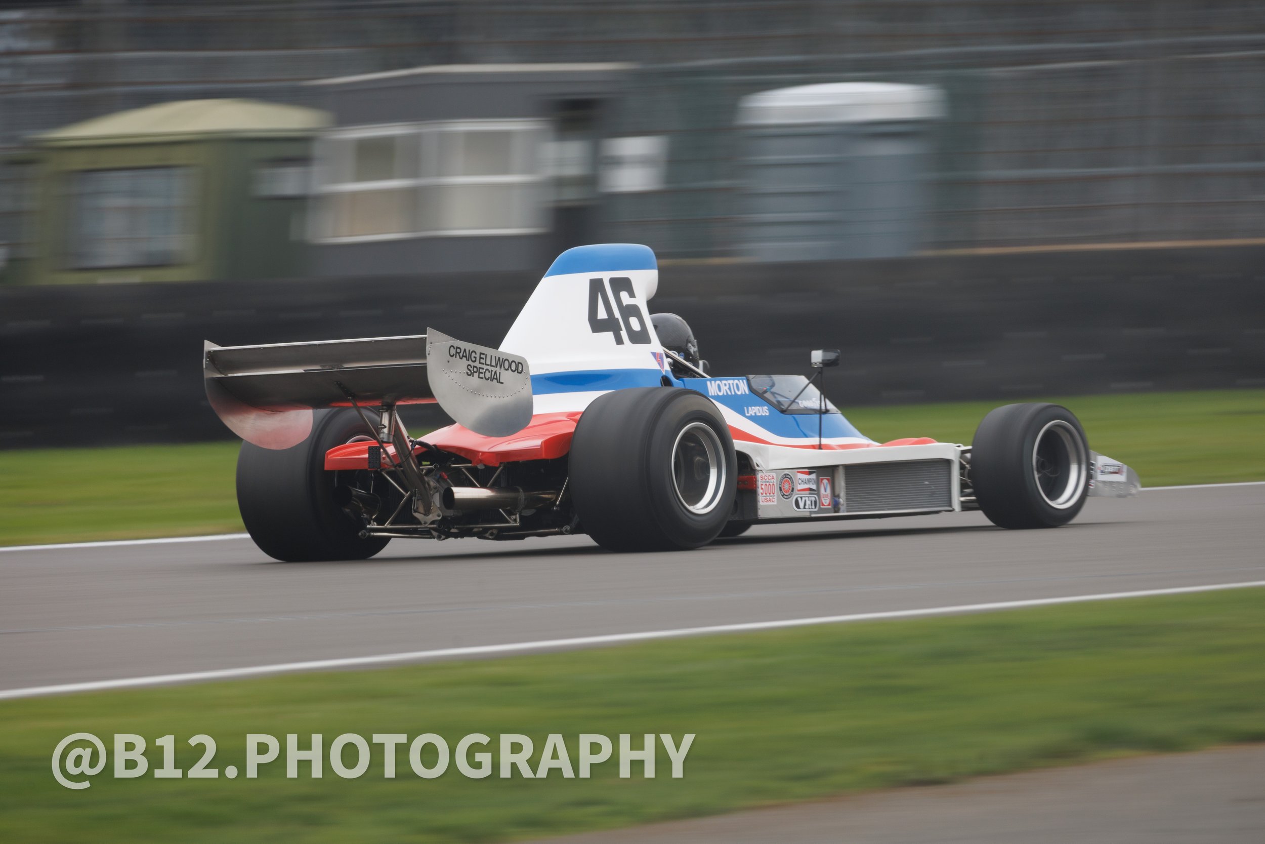 A vintage open-wheel race car speeding around a track with blurred background, featuring a white, blue, and red color scheme, large rear wing, and sponsor stickers.