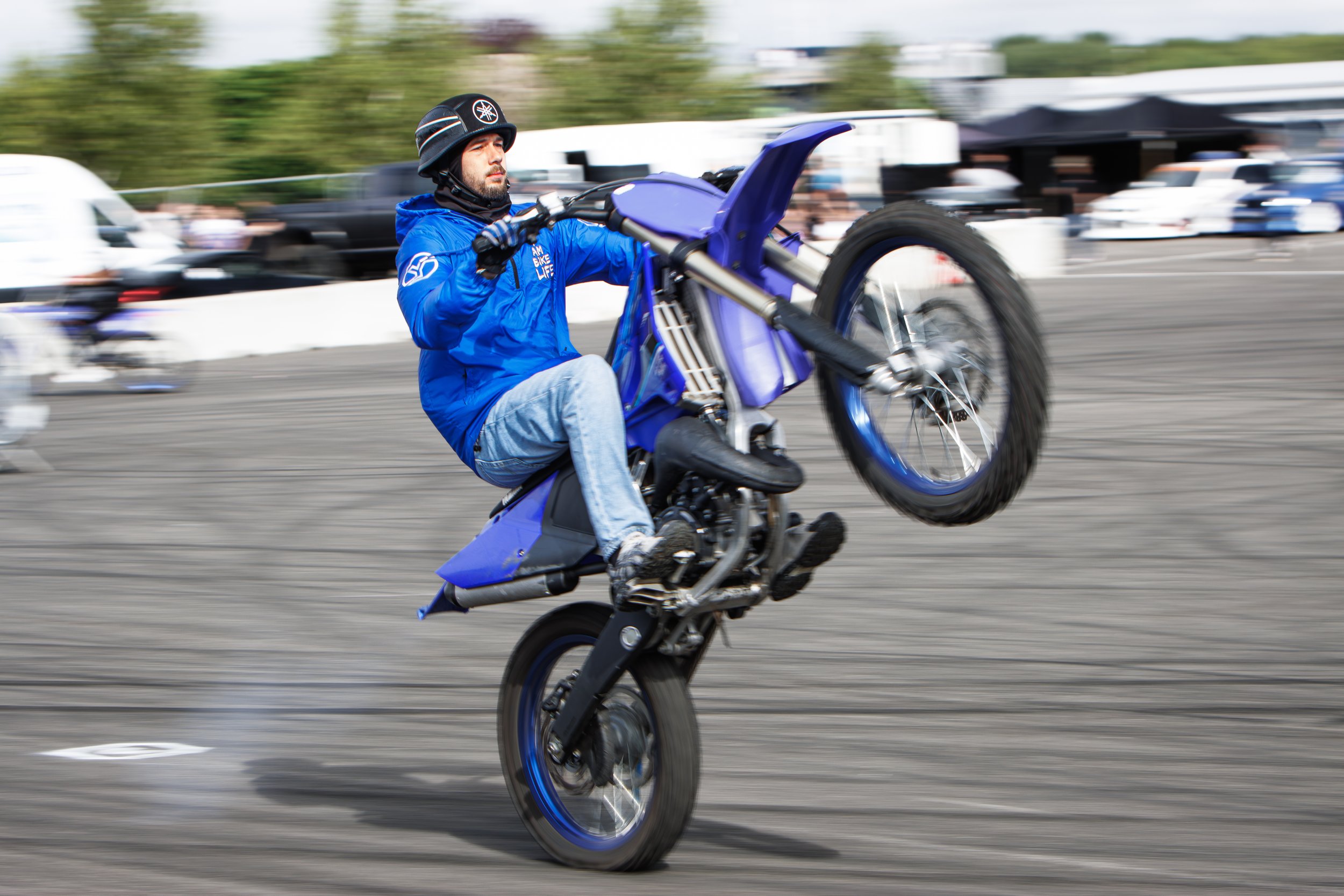 A man wearing a black helmet and blue jacket performing a wheelie on a blue motorcycle on a paved surface.