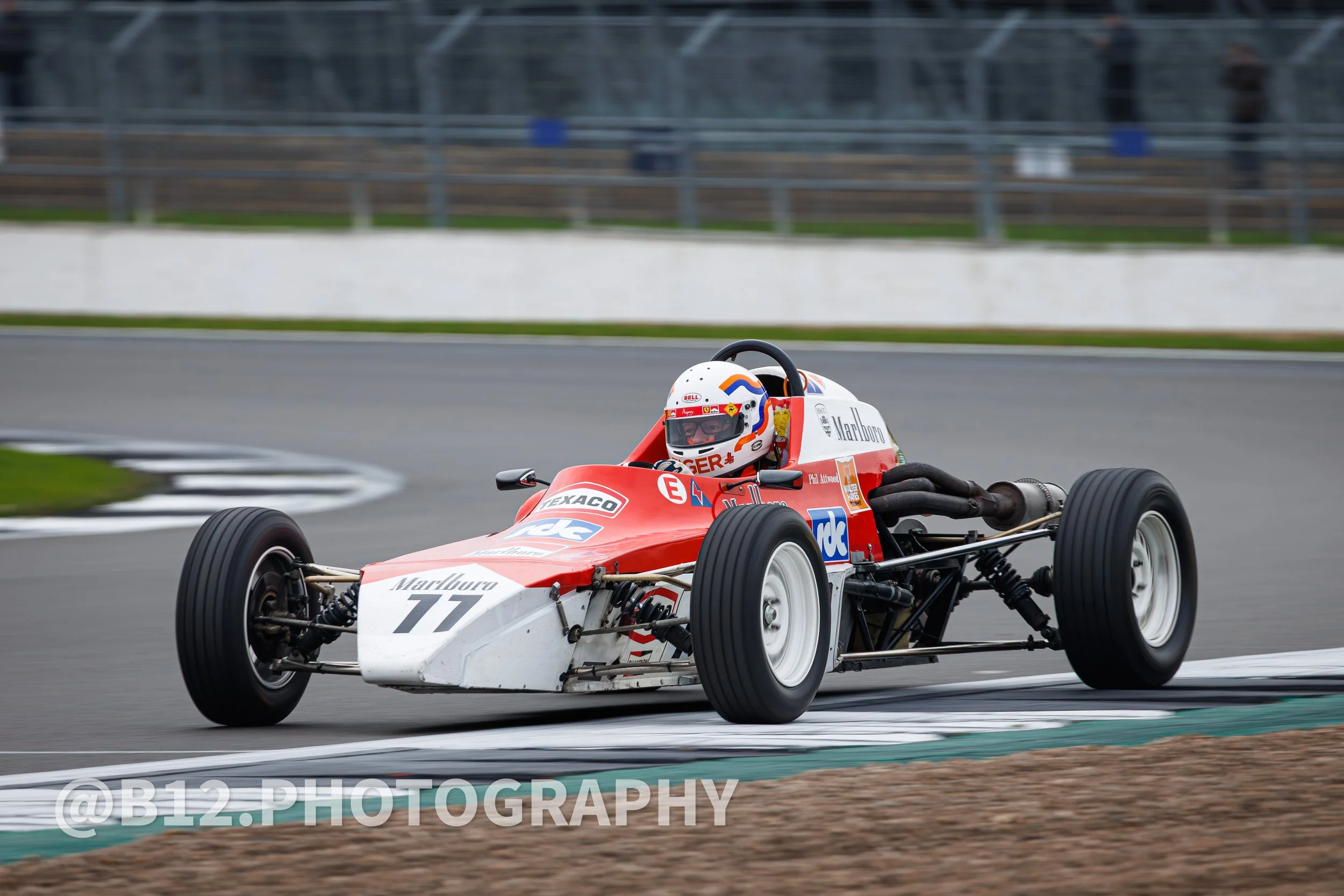 Vintage race car with the number 77 on the front, driven by a driver in a white helmet with red and blue details, navigating a turn on a racetrack.