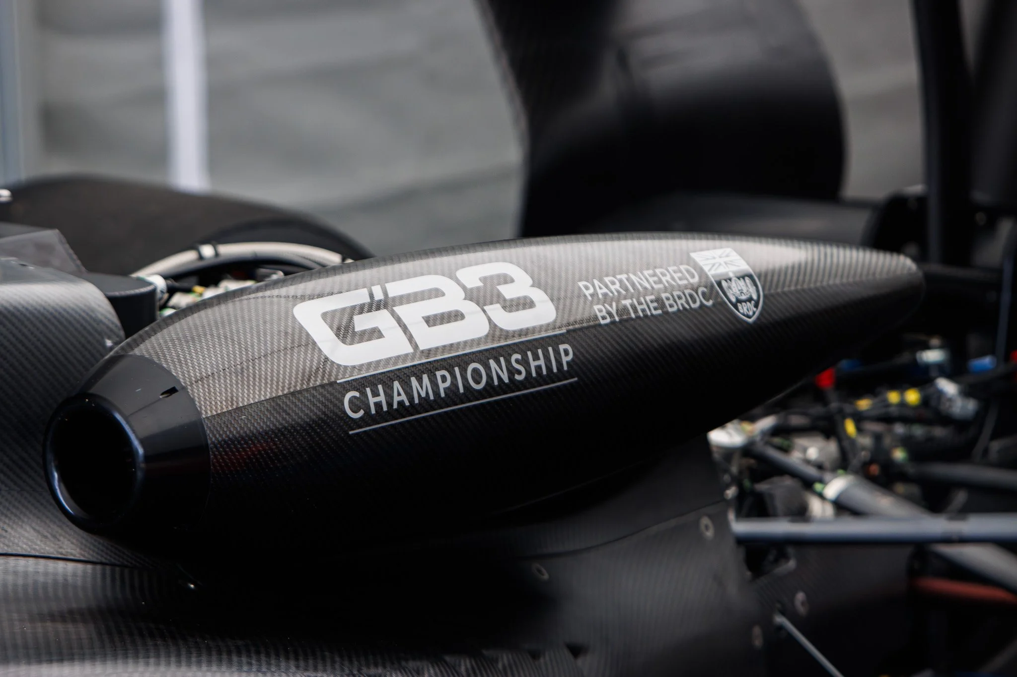 Close-up of a black carbon fiber Formula racing car part with white text reading "GB3 CHAMPIONSHIP PARTNERED BY THE BRDC" and a small logo. The background includes some wheels and mechanical components.