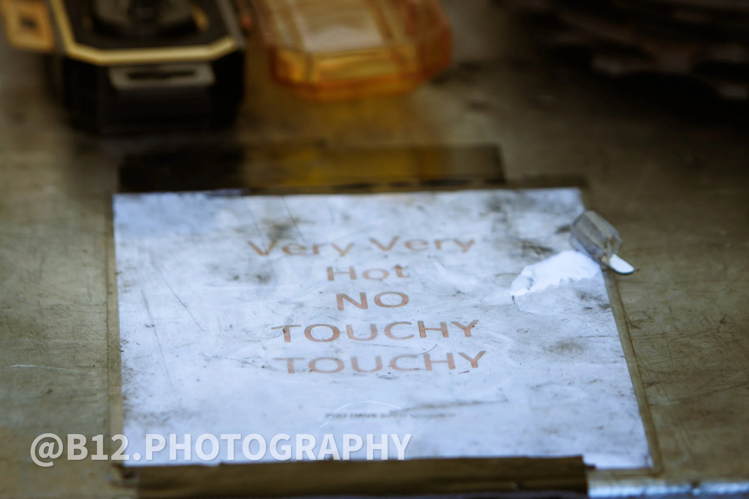 A sign that reads "Very Very Hot NO TOUCHY" in red letters on a reflective surface, placed on the floor. Items are visible in the background.