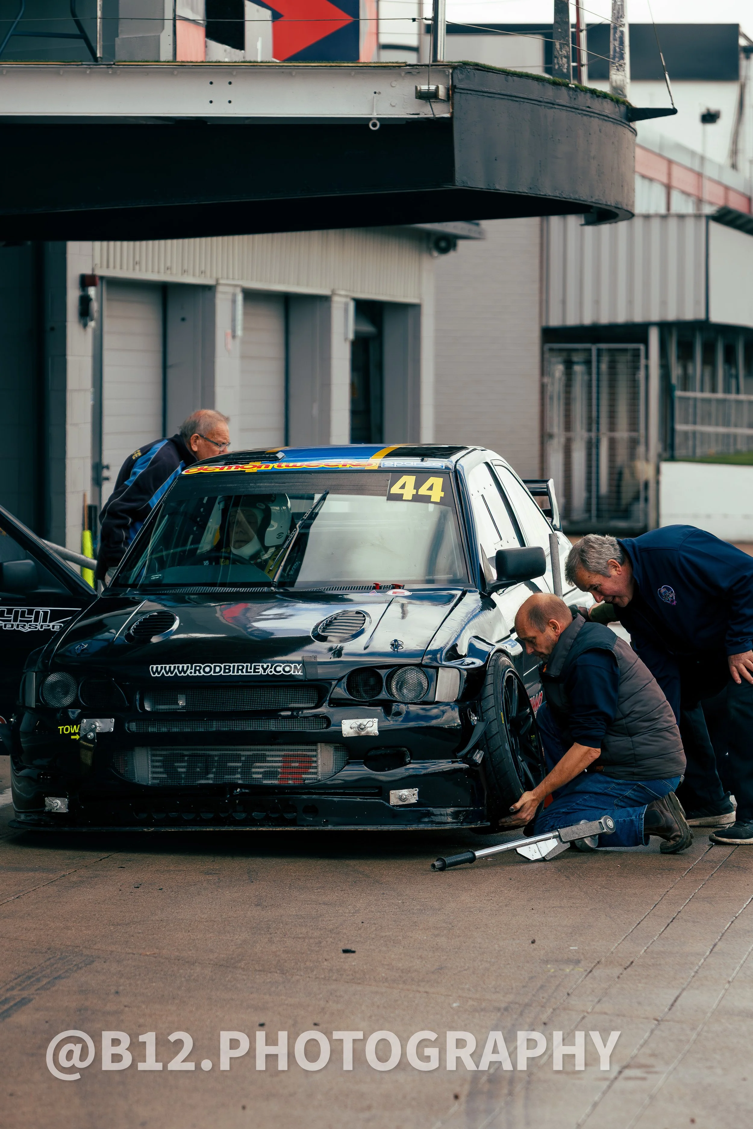 Three men working on a black race car in a garage area. The car has the number 44 on the windshield and a website URL on the hood. The men are inspecting or fixing the front left wheel.