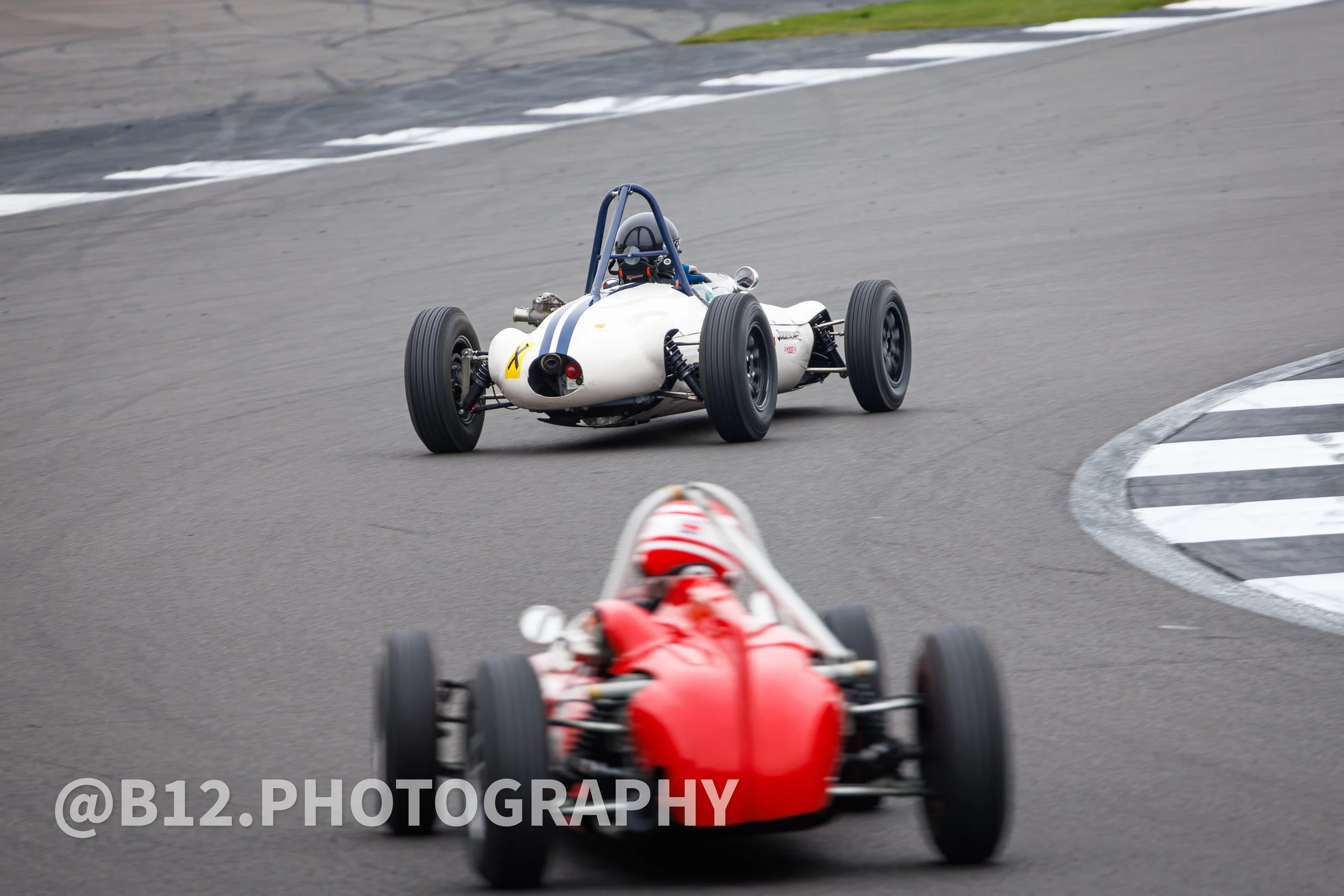 Two vintage open-wheel race cars on a racetrack, one red in the foreground and one white behind, navigating a turn.