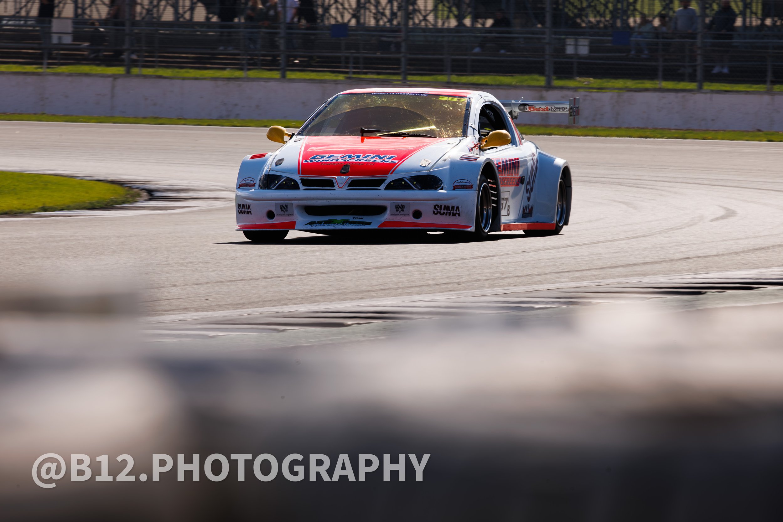 Race car on a race track with a blurred foreground and a chain-link fence in the background.