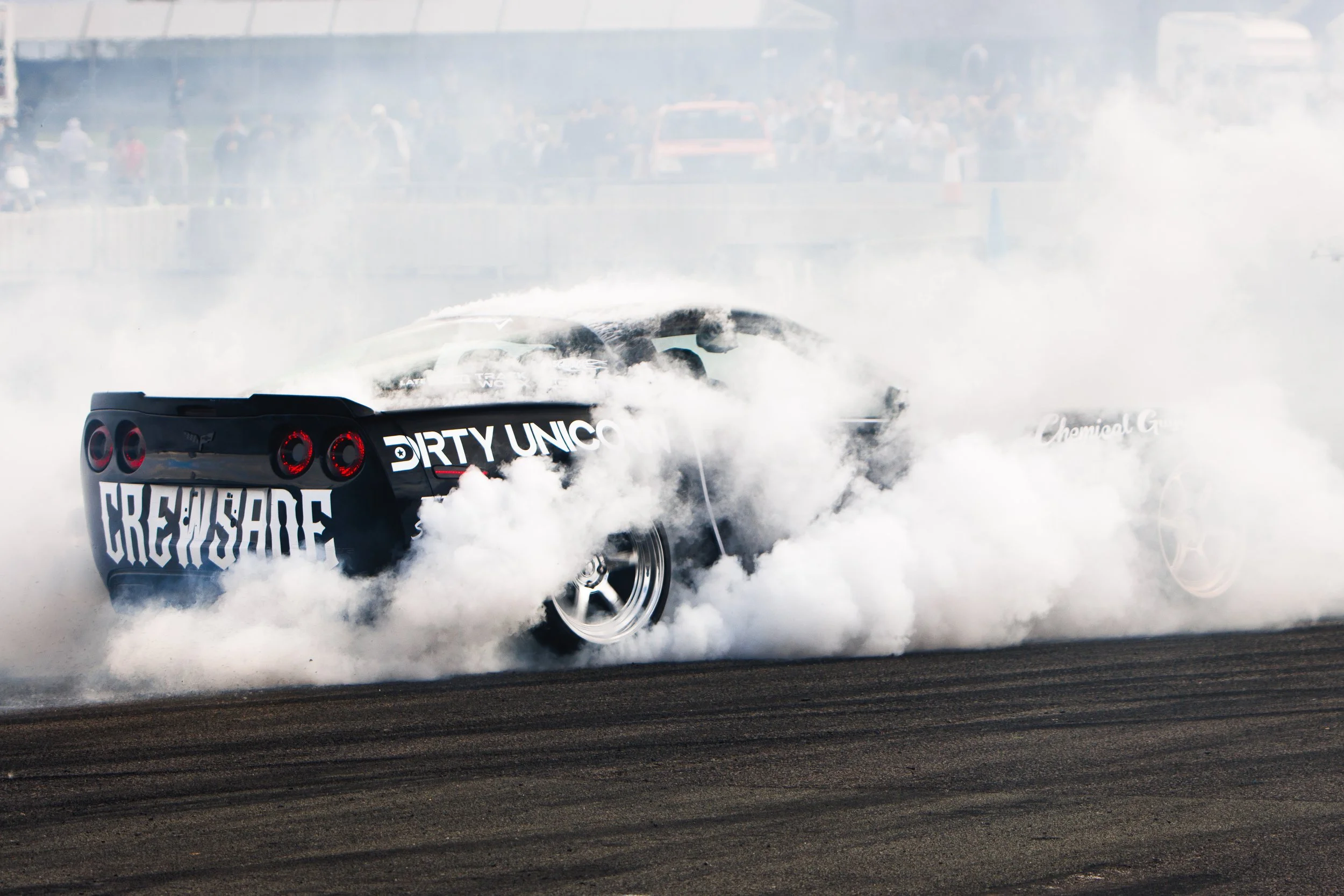 A black racing car performing a burnout on a track, creating thick white smoke around it. The car has racing tires and is surrounded by spectators.