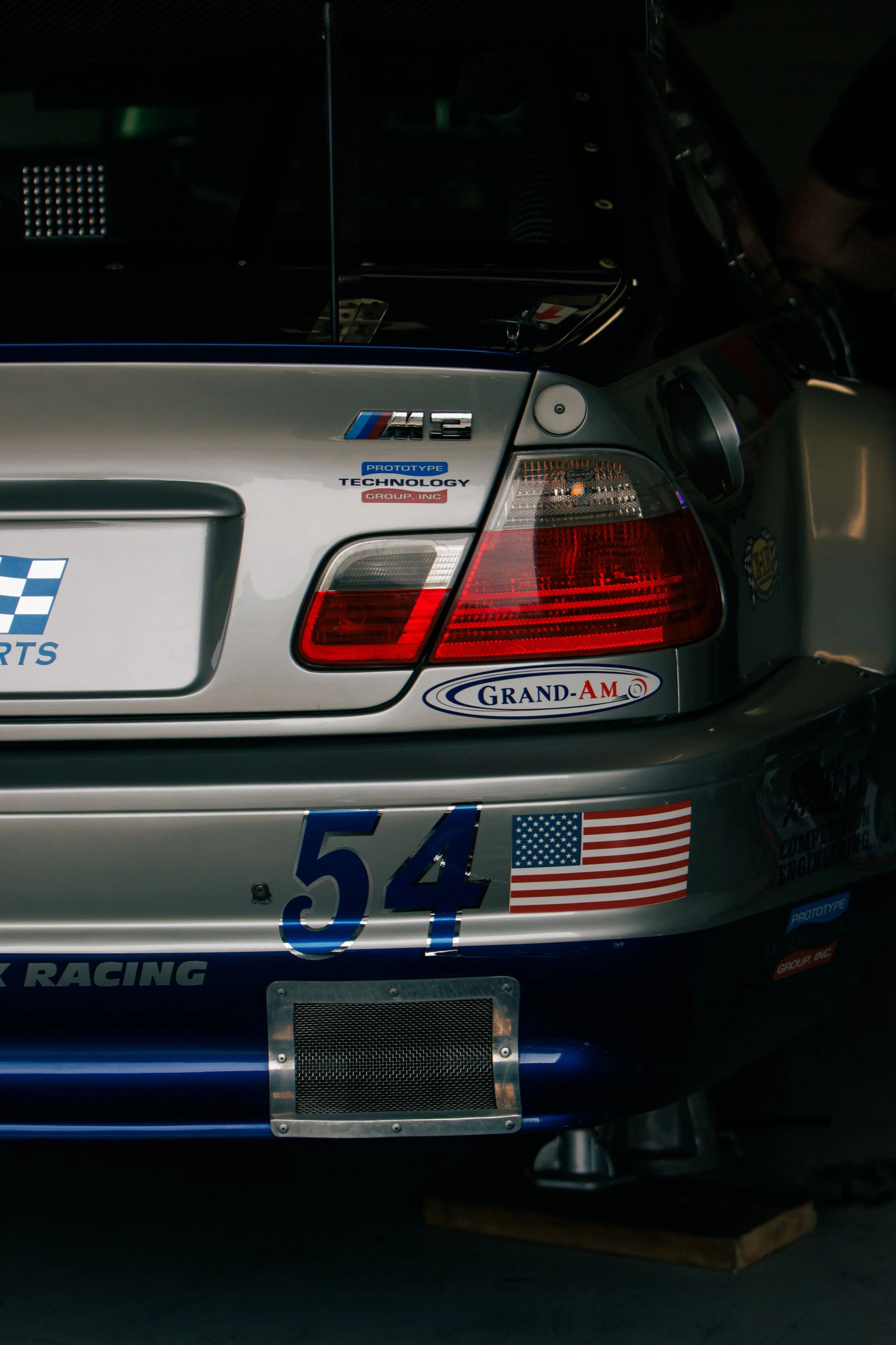 Back view of a race car with various sponsor decals, including an American flag, number 54, and BMW M3 badge, in a dimly lit garage.