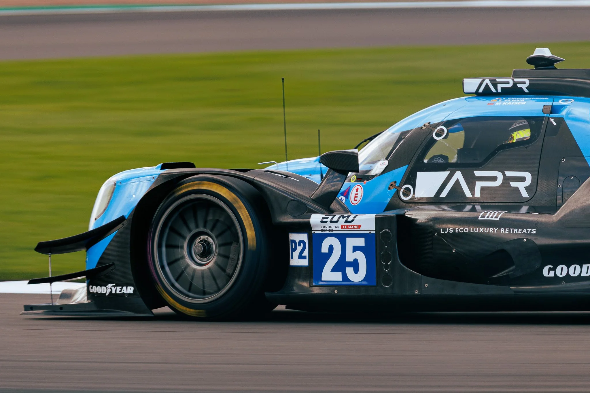 A blue and black prototype race car with the number 25 on the side, racing on a track with blurred grass background, featuring Goodyear tires and various sponsor logos.