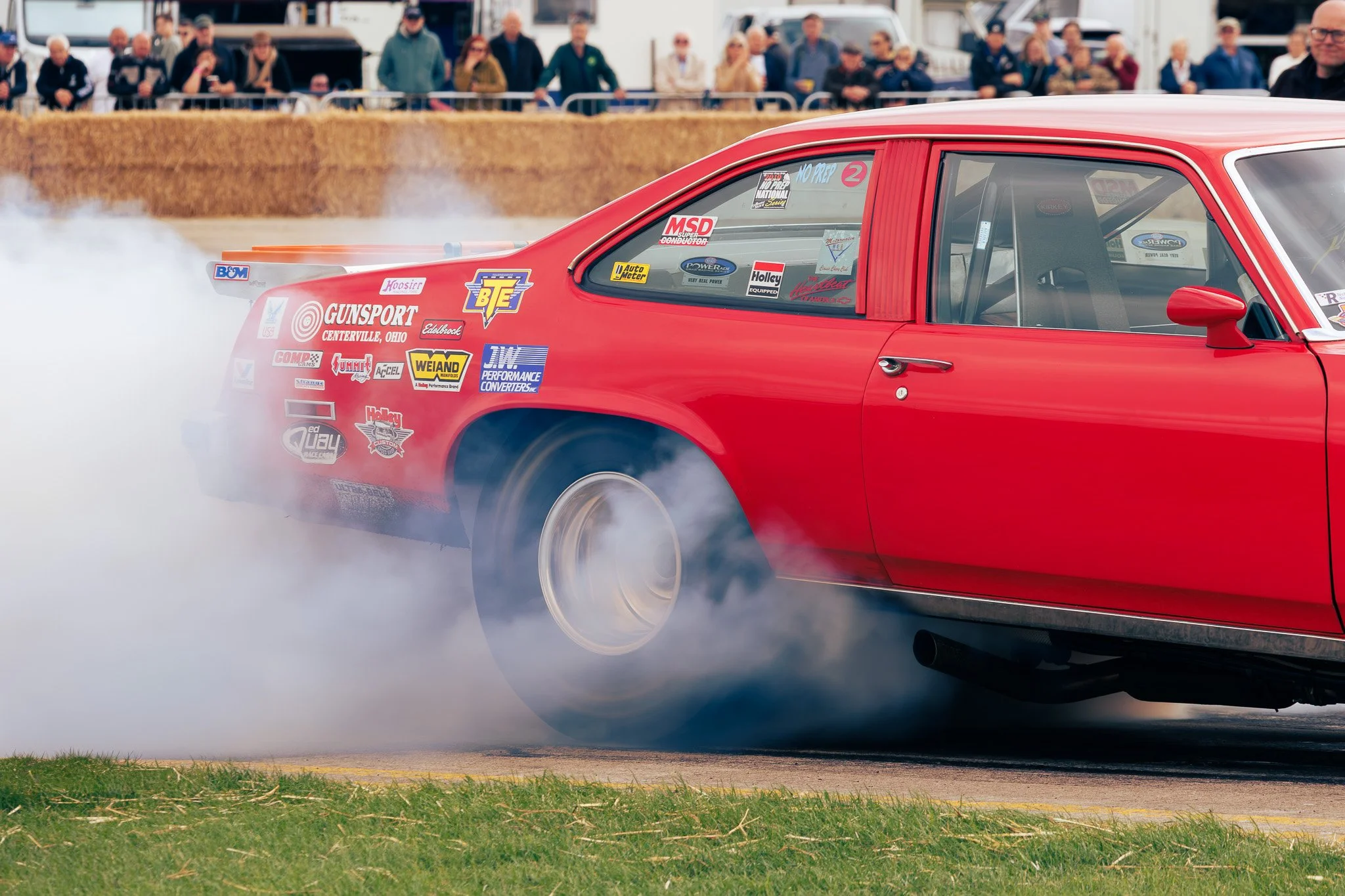 Red race car performing a burnout on a track, smoke billowing from the rear tires, with spectators in the background.