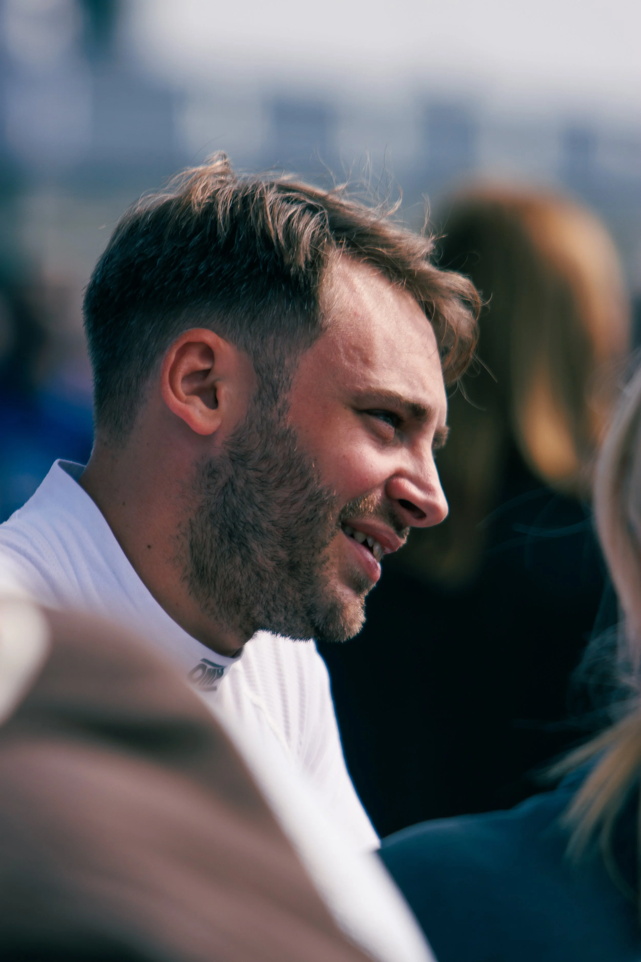A smiling man with short brown hair and a beard, wearing a white shirt, appears to be engaged in conversation.