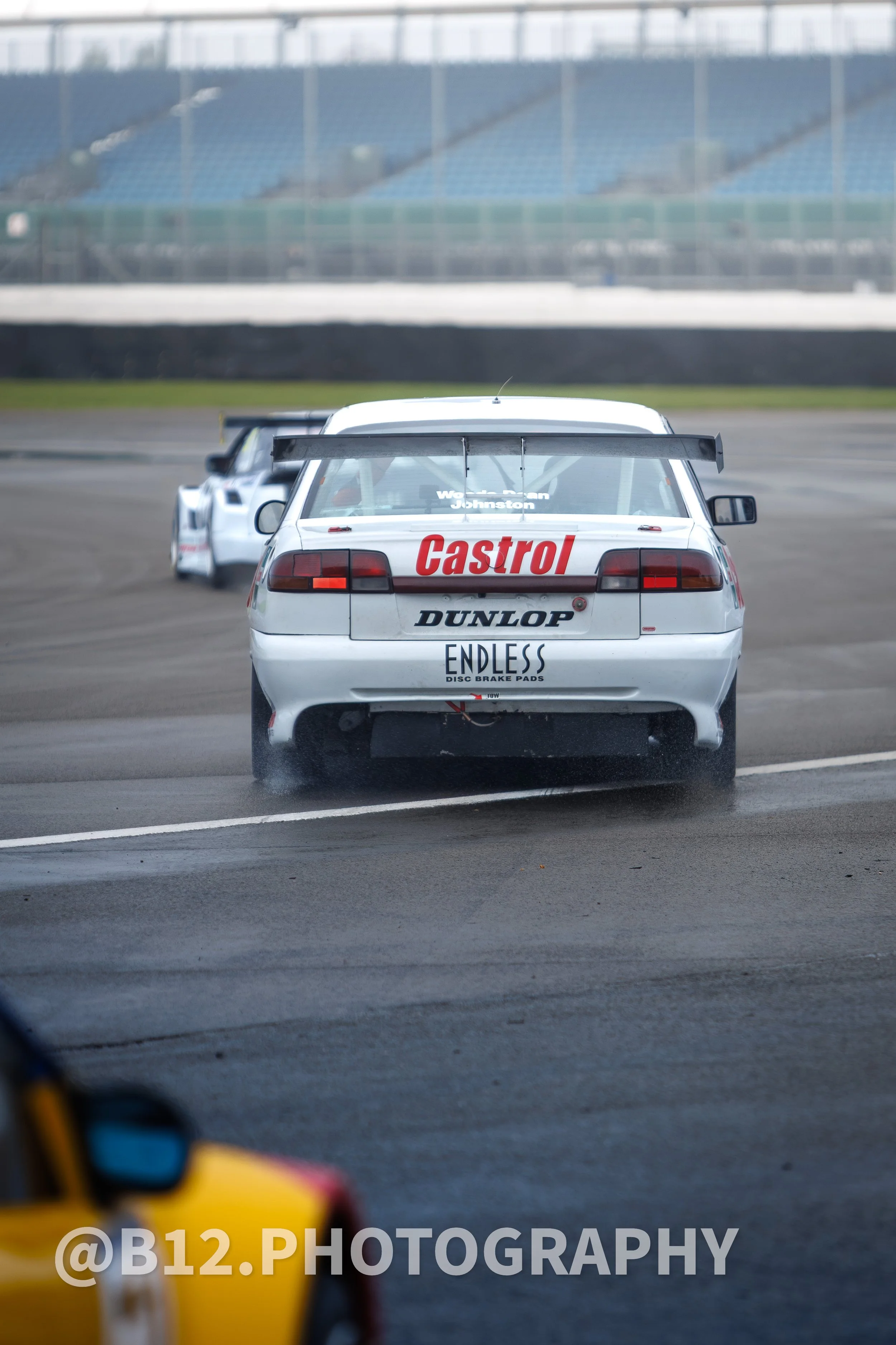 Two race cars on a track, with the car in the foreground featuring sponsor logos 'Castrol,' 'Dunlop,' and 'Endless.' The cars are moving, leaving tire marks behind, with a grandstand in the background.