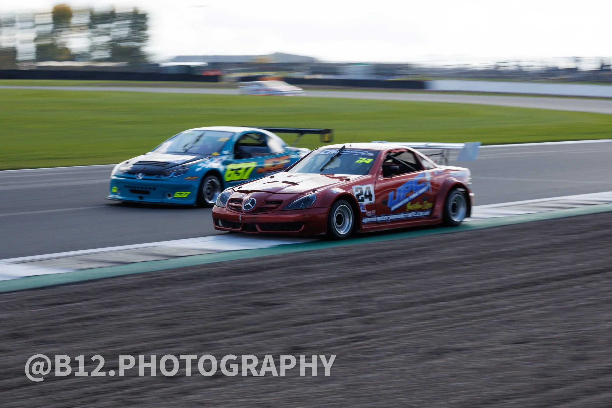Two race cars, a red one and a blue one, racing on a track, with the red car in the lead and the blue car close behind, both speeding around a curve.