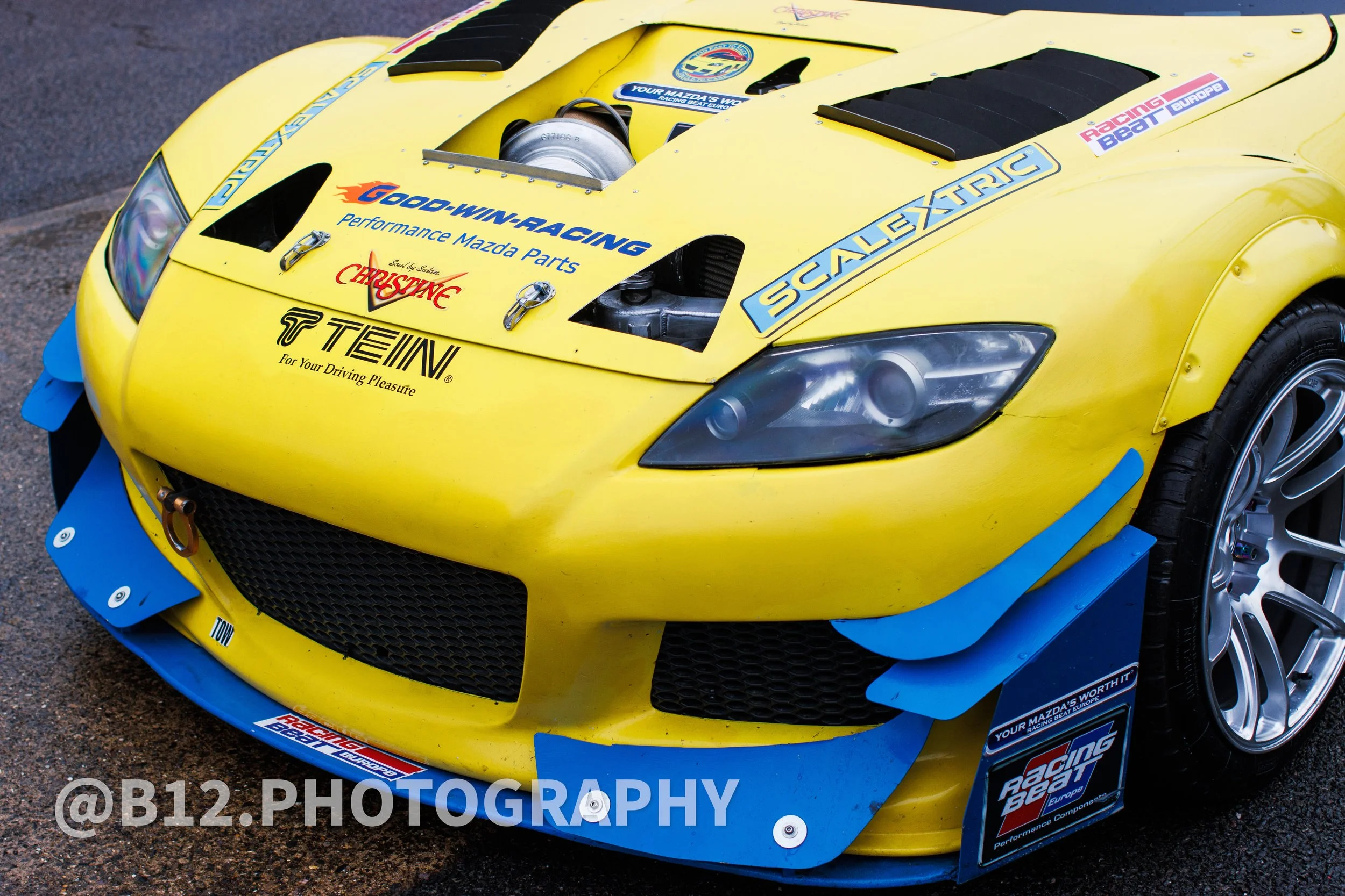 Yellow race car with aerodynamic blue splitters, black vents, and decals including 'GOO-WIN-RACING,' 'SCALA ELECTRIC,' and 'Racing Beat Europe,' parked on asphalt.