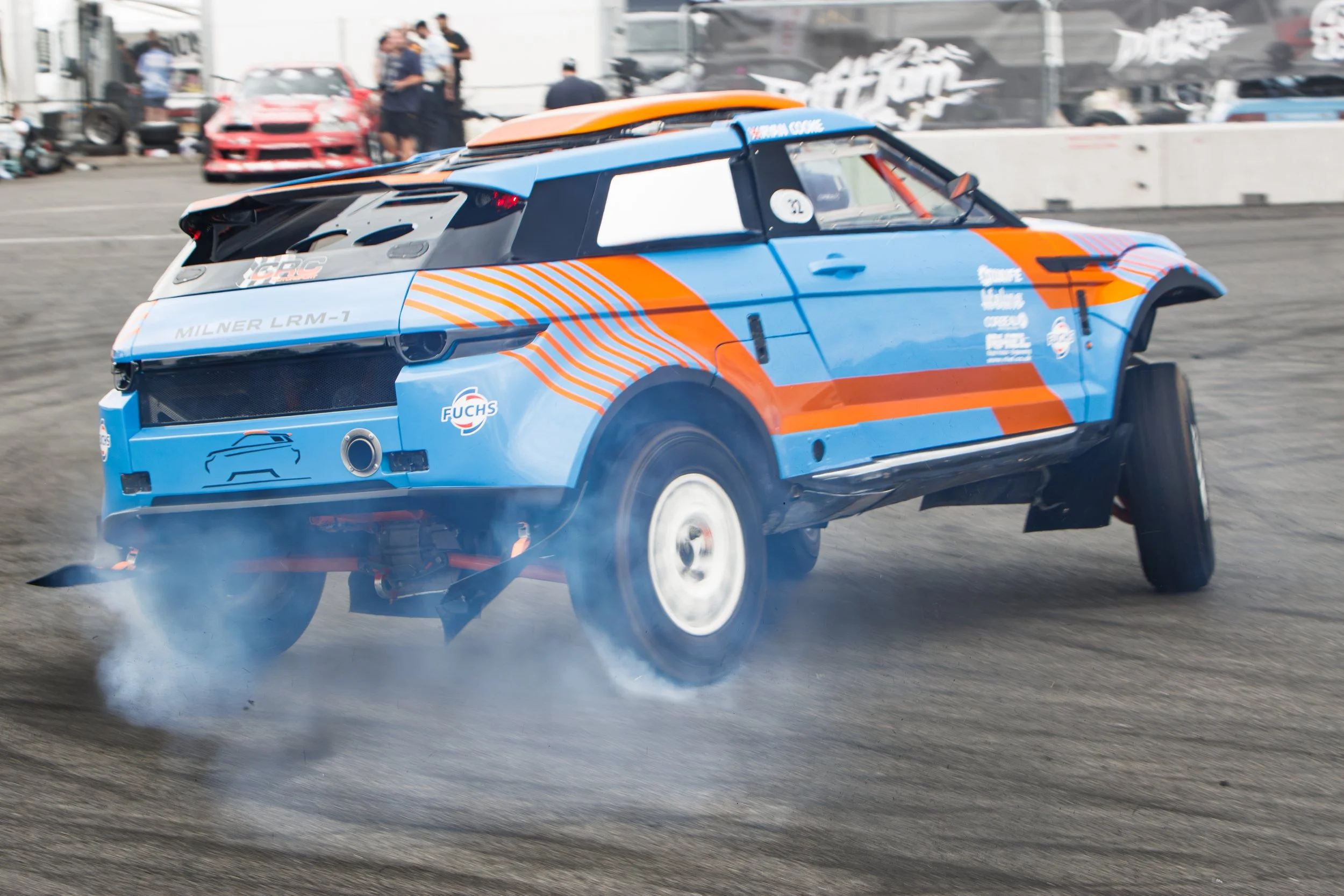 Race car drifting on a track with smoke coming from the tires, surrounded by other vehicles and people in the background.