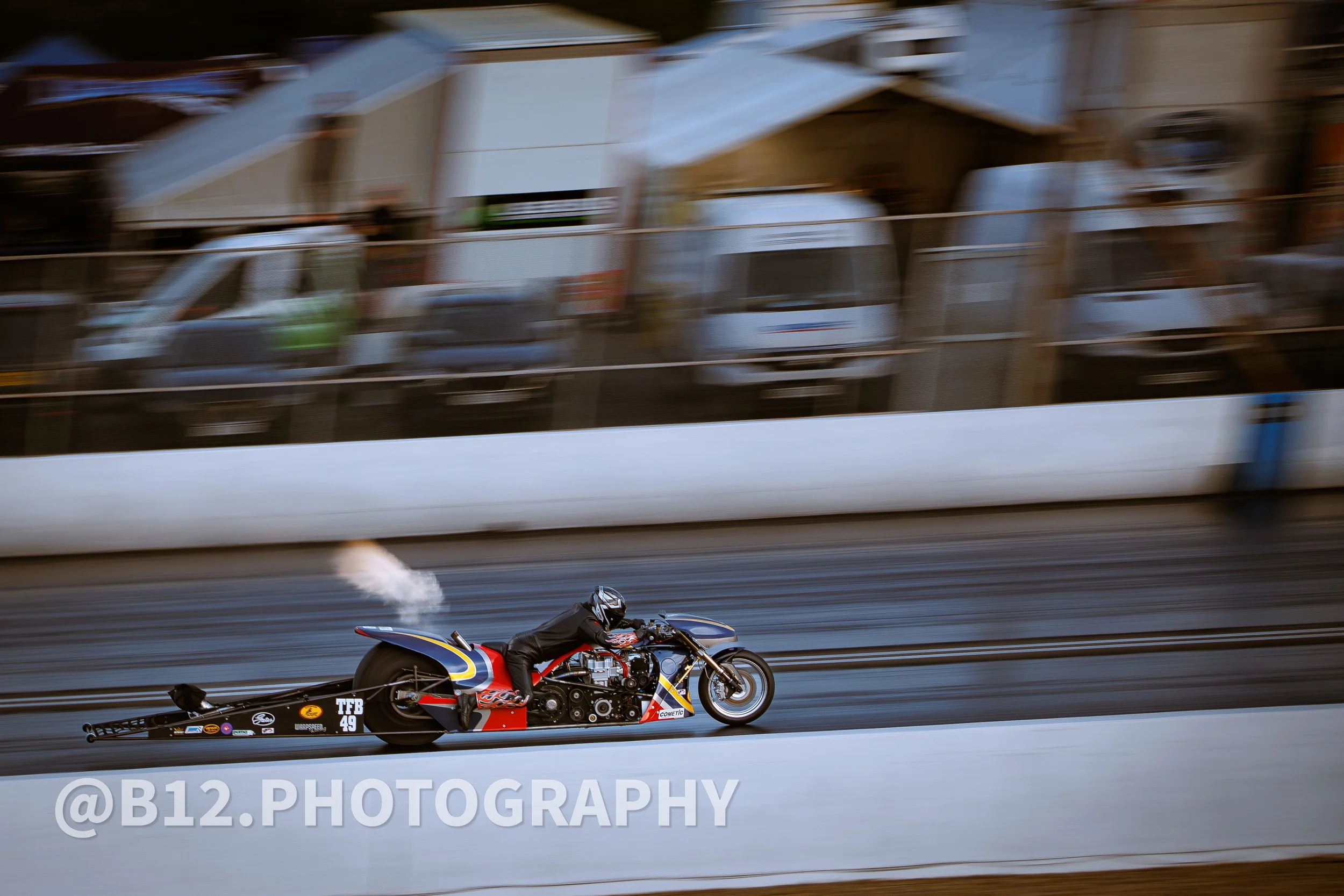A motorcycle drag racing on a track with a rider wearing a helmet and black racing suit. The motorcycle is a top-fuel bike with a long body and large rear tire, emitting smoke from its exhaust. The background is blurred, showing trucks and trailers.