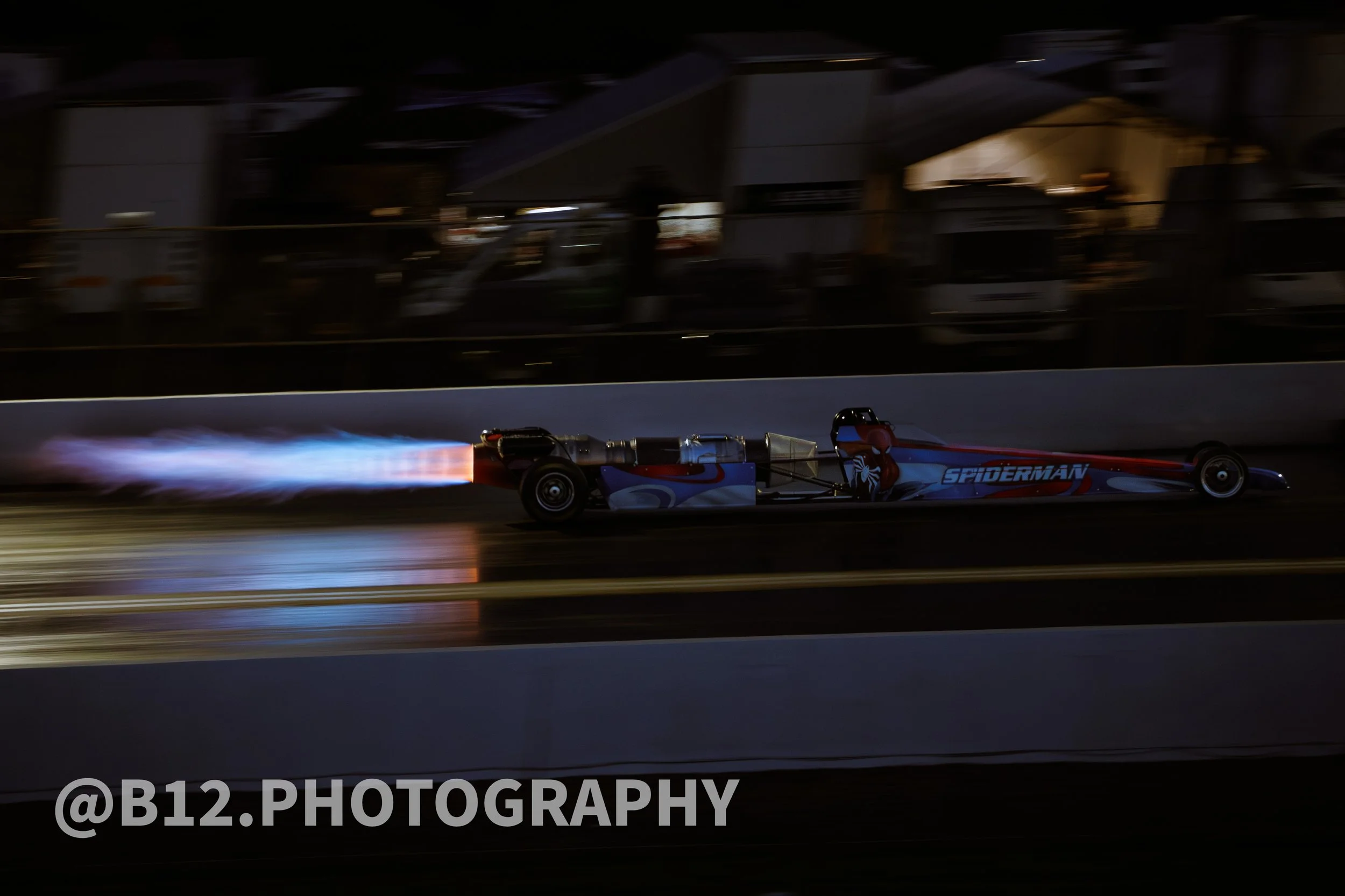 A drag race car with Spider-Man themed paint job speeding at night, producing flames from the exhaust, on a drag strip with blurred background and track markings.