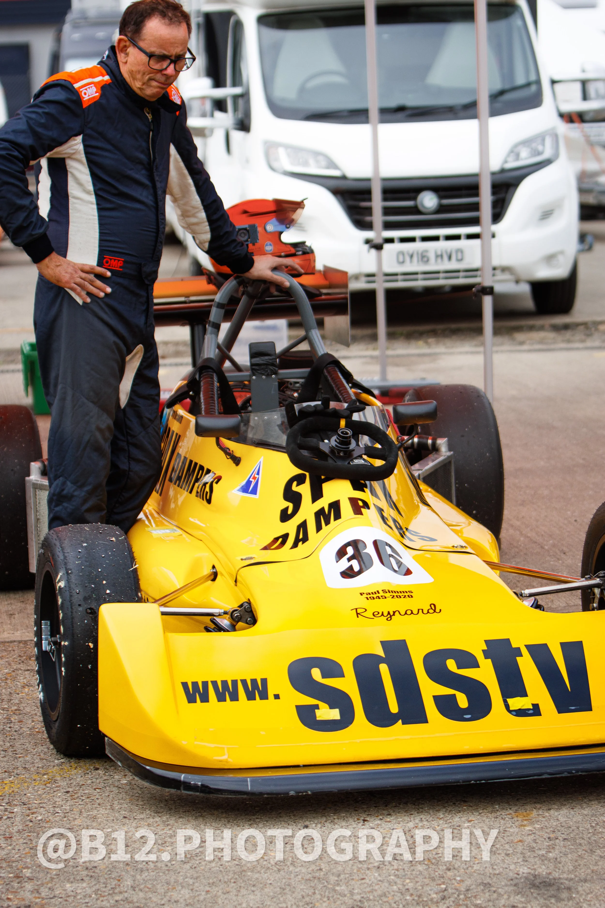 A man in a racing suit standing next to a yellow racing car with the website www.sdsps.com on the front. The man is looking down at the car, in a paddock area with trucks and cars in the background.